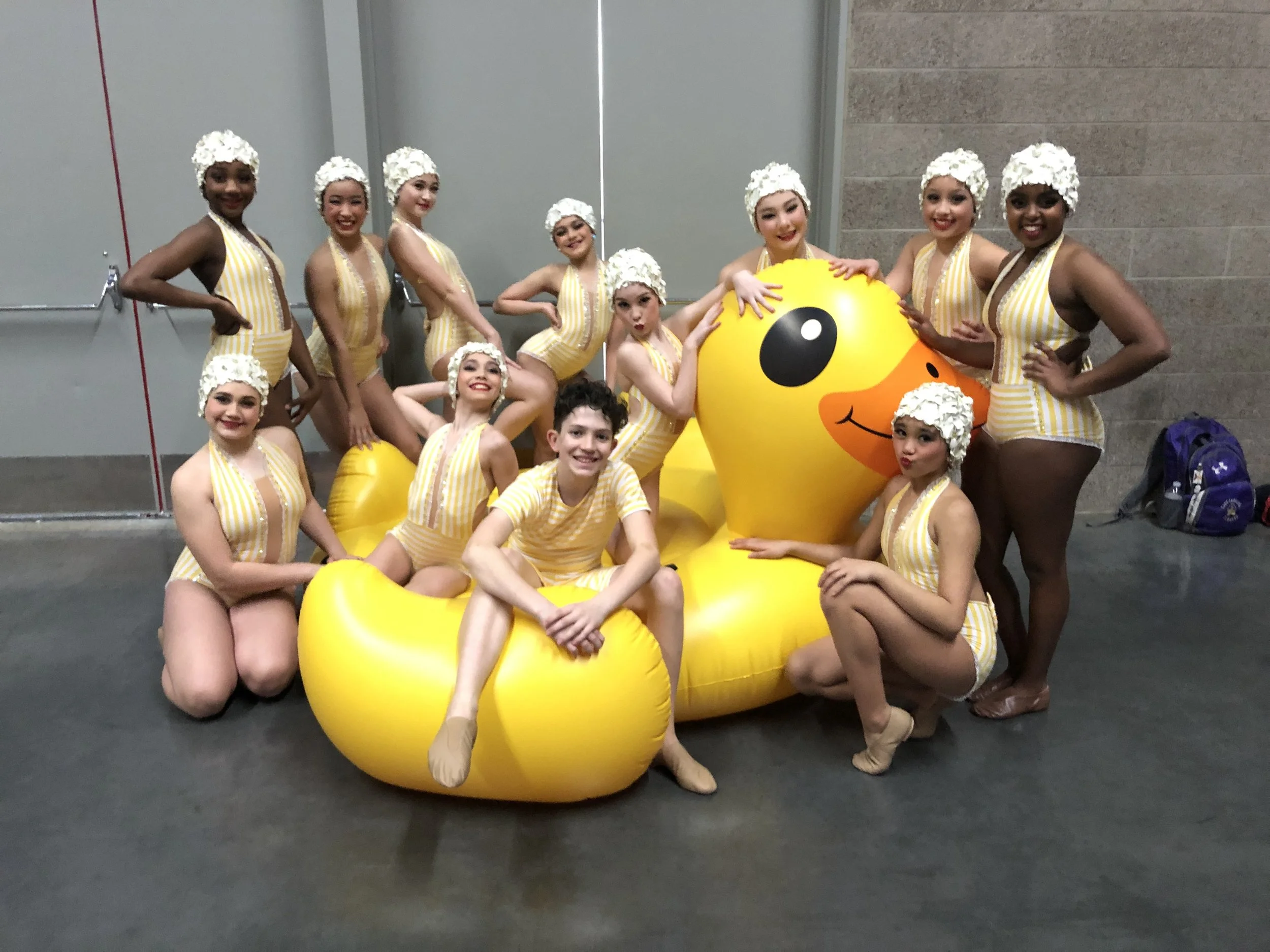 Group of smiling young girls in yellow and white striped costumes with white headpieces, posing with large yellow duck float.