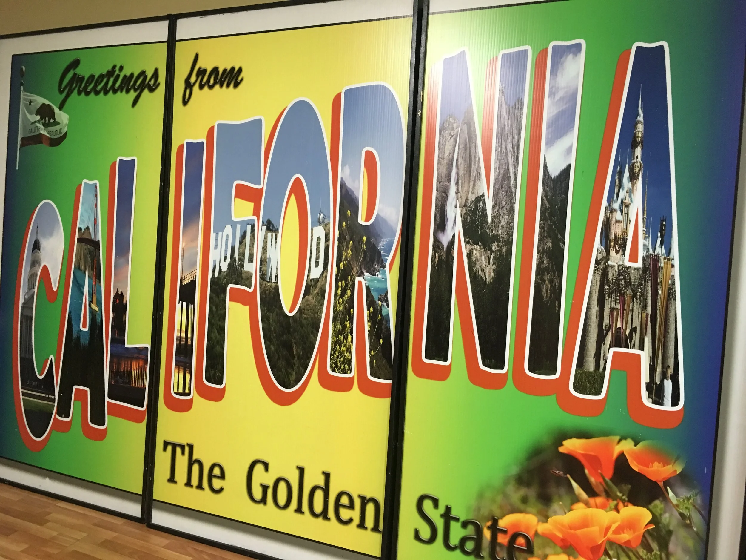 Colorful welcome sign with the text 'Greetings from the Golden State' and images of California landmarks and scenery, including mountains, a lighthouse, and orange flowers.