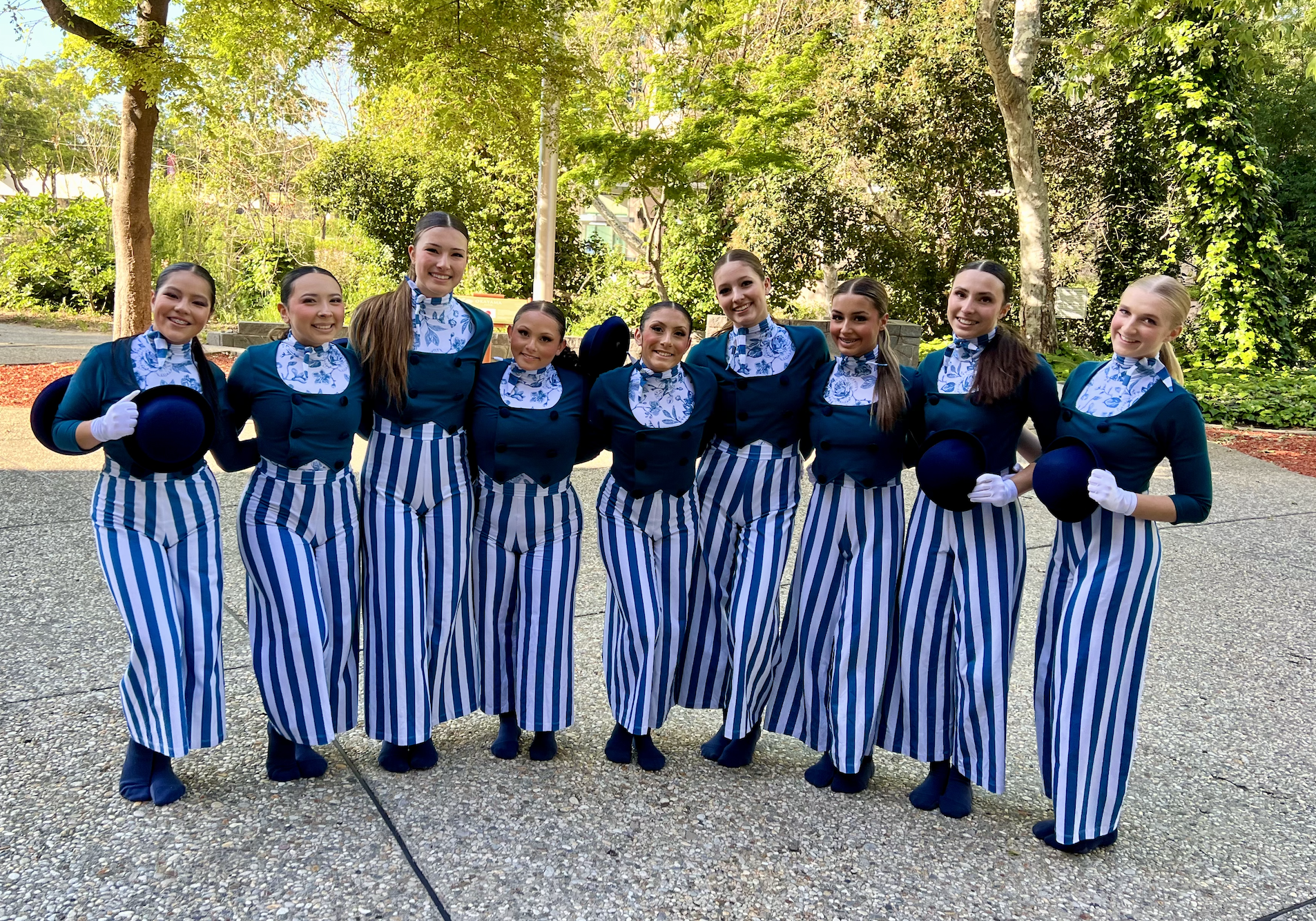 A group of nine women dressed in blue and white striped pants, navy blue jackets, and matching scarf neckties, standing outdoors on a sidewalk surrounded by green trees, posing for a photo with smiles and some holding black hats and wearing white glo