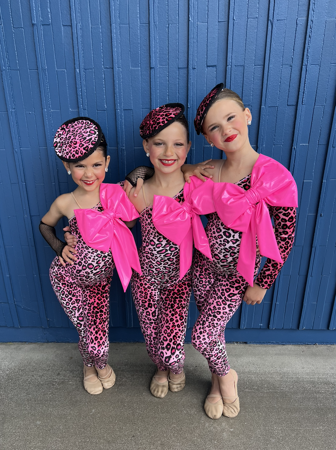 Three young girls dressed in matching pink and black leopard print costumes with large pink bows, leopard print hats, and beige dance shoes, standing in front of a blue wooden wall.
