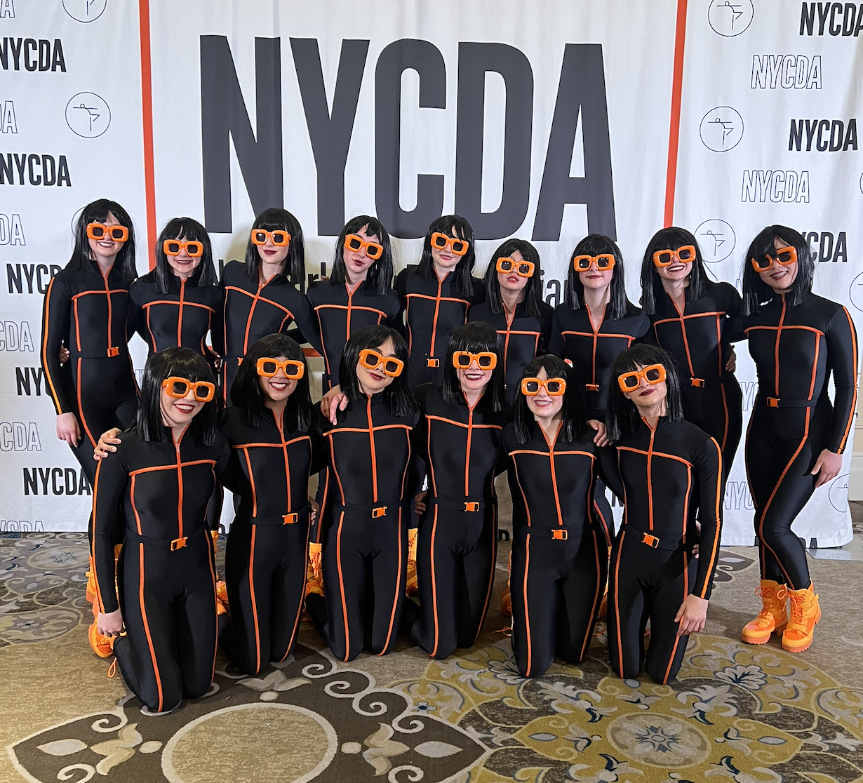 A group of women dressed in black jumpsuits with orange accents, wearing orange glasses, standing in front of a NYCDAY backdrop at an event.
