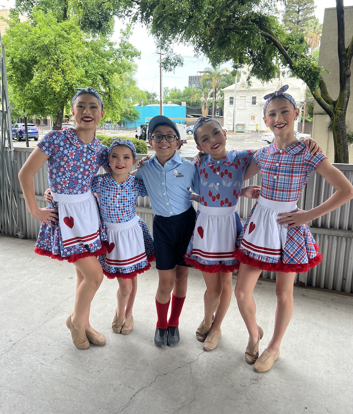 Group of five children dressed in vintage-inspired outfits, standing outdoors on a concrete patio with green trees and buildings in the background. They are smiling and have their arms around each other.