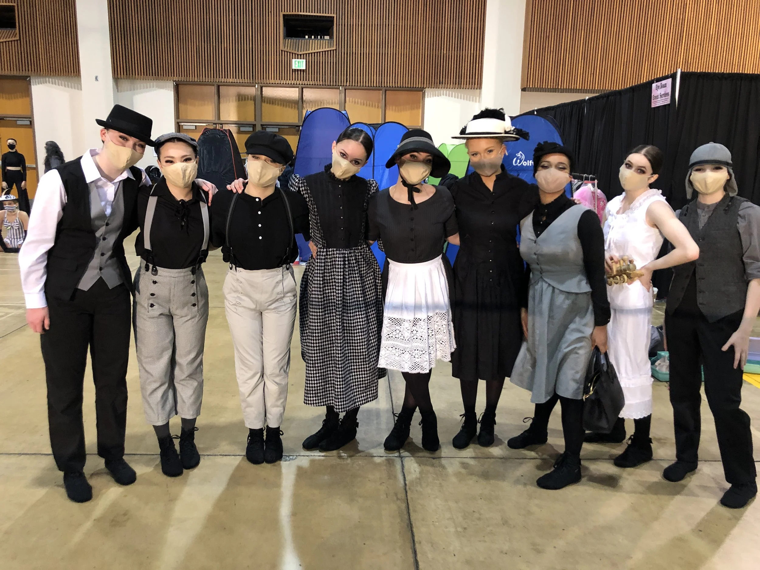 Group of nine women dressed in vintage or historical costumes, wearing masks, standing indoors in front of colored tents and black curtains.
