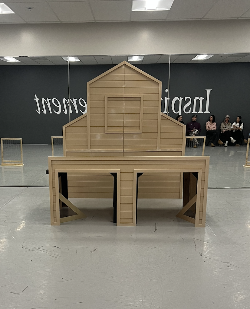 A large wooden barn-shaped display structure in a room with a mirror and people sitting against the wall in the background.