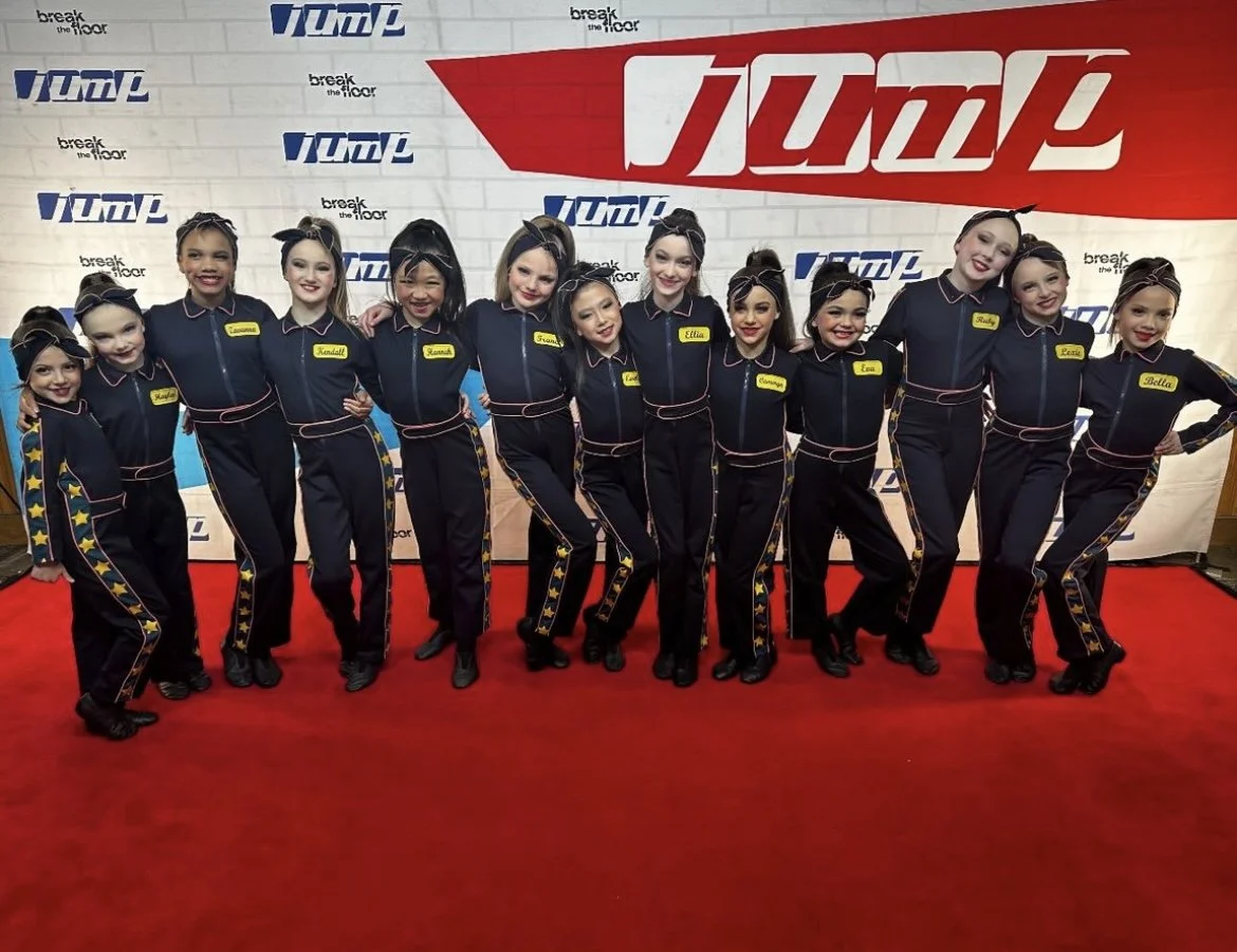 A group of young girls in matching navy blue dance costumes with star details on the sides, posing together on a red carpet at a performance event. They are smiling and standing with their arms around each other, in front of a backdrop with the words
