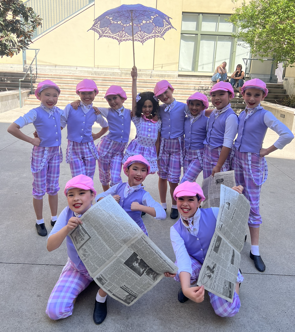 A group of young children dressed in 1920s-style costumes, including pink berets, purple vests, and checkered pants or skirts, pose outdoors with a woman wearing a vintage dress. They are smiling and holding newspapers. One child is holding a purple 