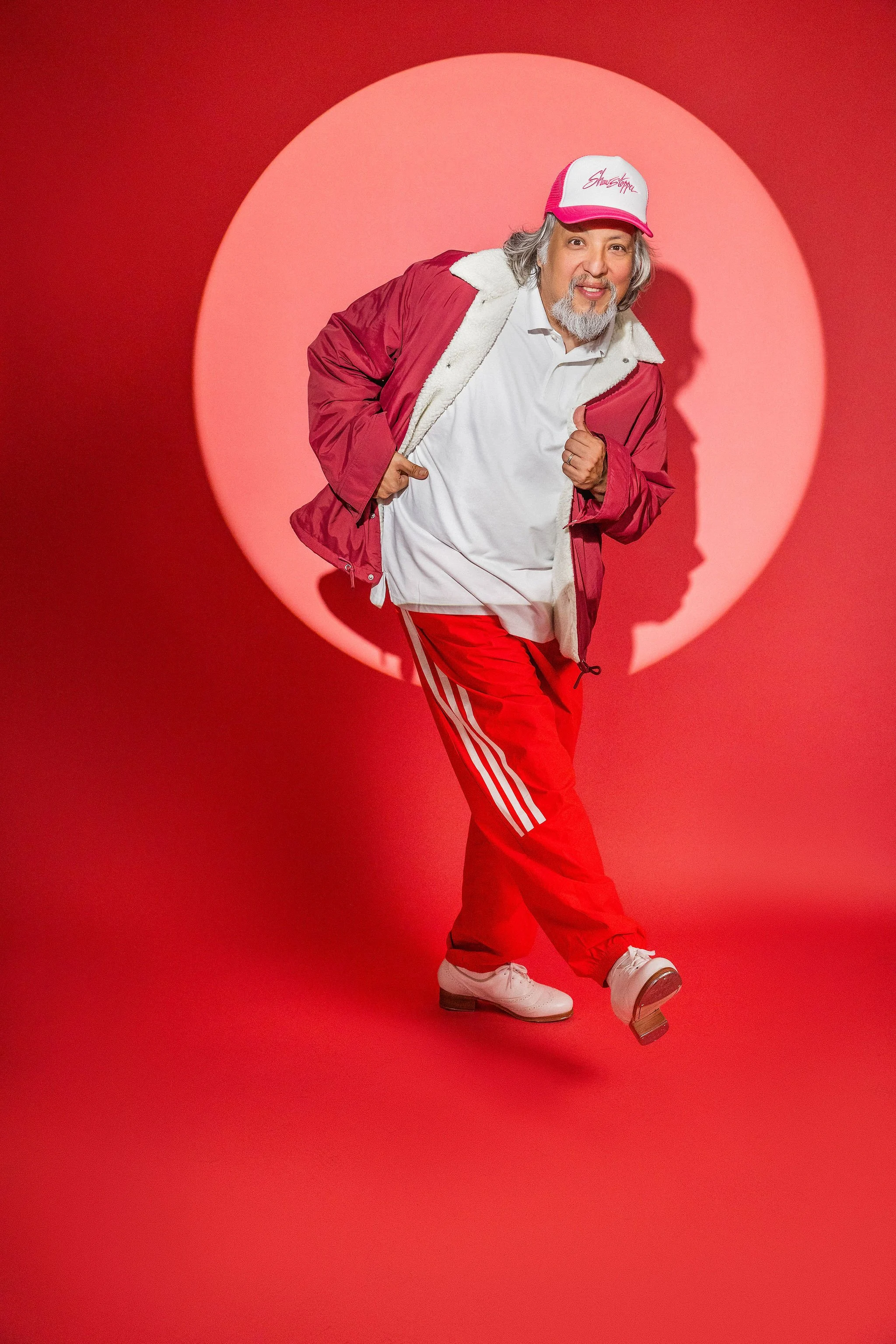 A man with gray hair and beard wearing a white shirt, red jacket with white lining, red pants with white stripes, and white shoes. He is also wearing a white and pink cap with red writing on it, standing against a red background with a spotlight creating a circular light effect.