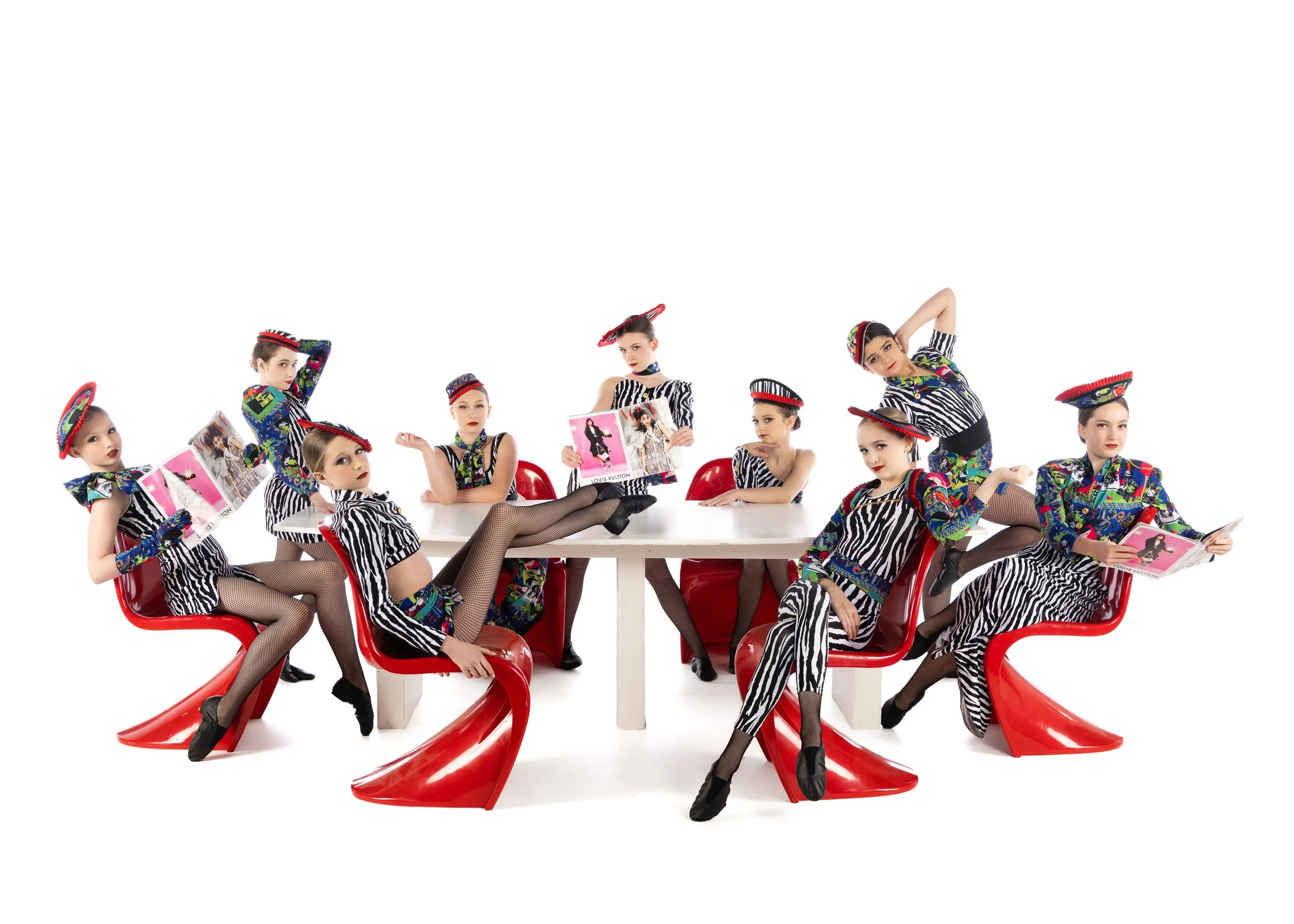 Group of women dressed in colorful, zebra-striped and vibrant patterned clothing, sitting and standing around a white table with red chairs, holding fashion magazines, and wearing matching red hats.