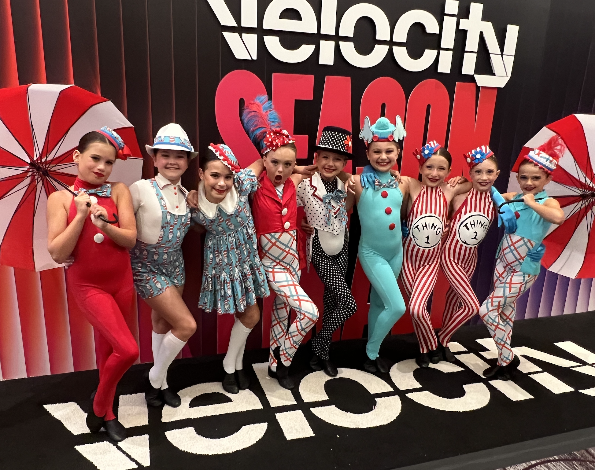 A group of children dressed in colorful costumes from the book and movie 'The Cat in the Hat', standing together in front of a large sign that reads 'Welcome'. Some children are holding red and white umbrellas.