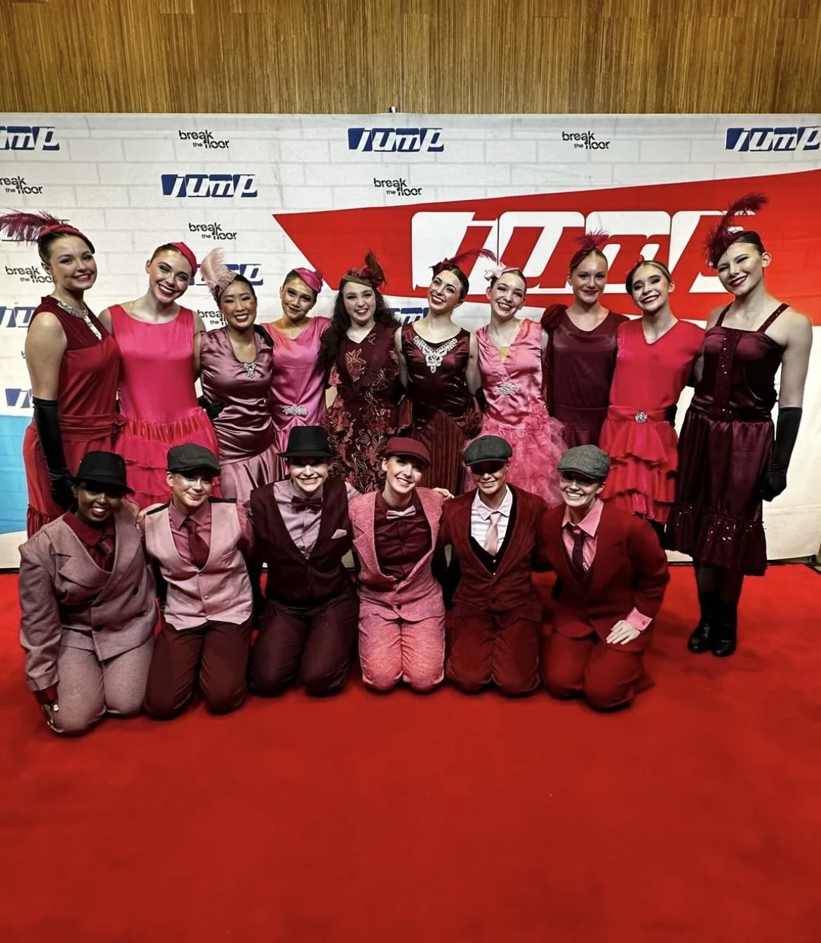 Group of performers, women and men, dressed in vintage-style costumes with shades of red, pink, and black, posing on a red carpet in front of a backdrop with logos.
