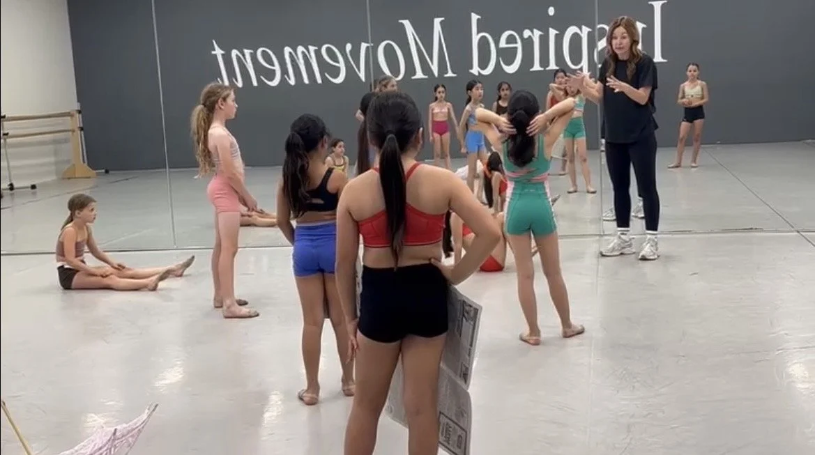 Group of young girls in dance costumes in a studio, listening to an instructor with a mirror and ballet barres in the background.