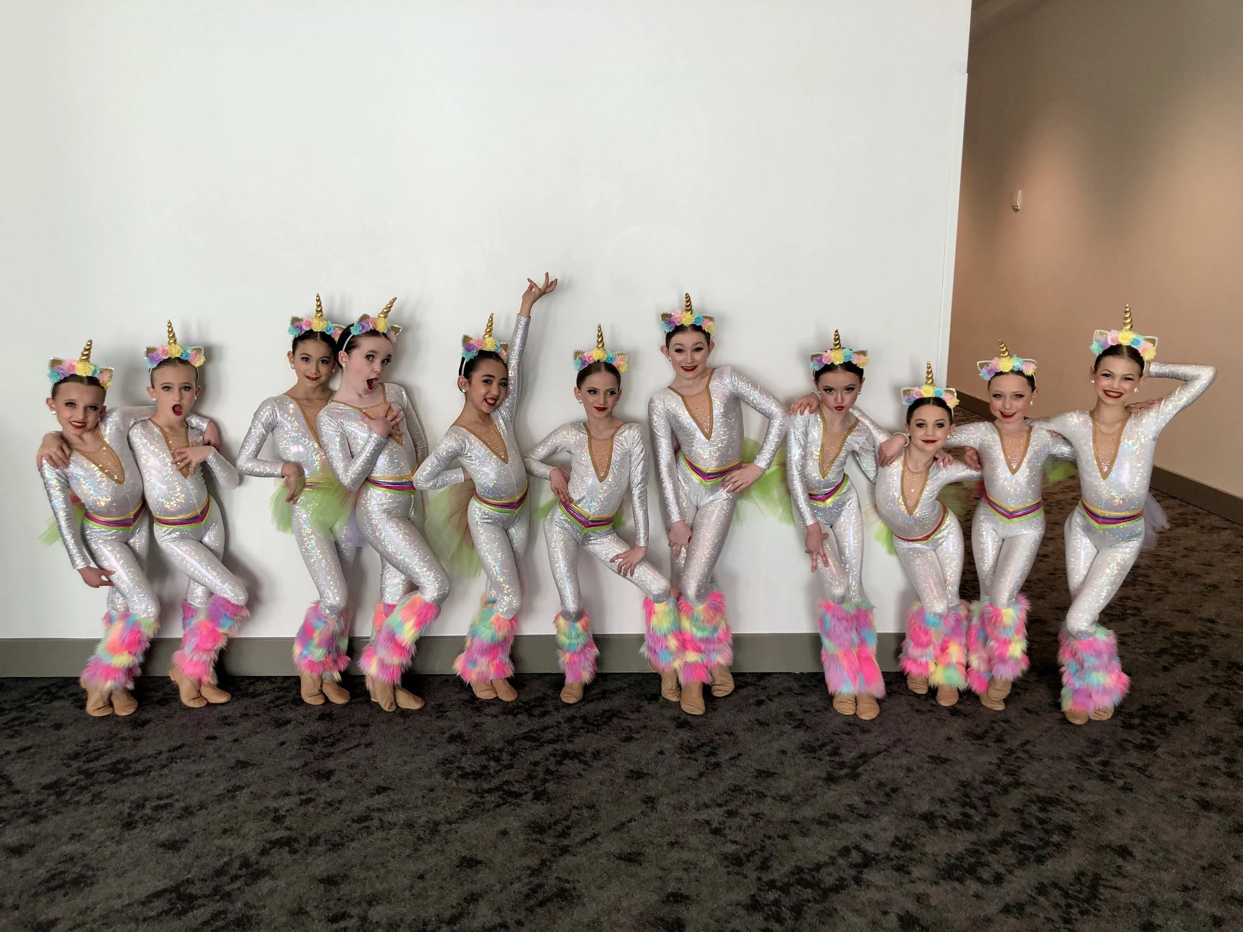 A group of young girls dressed in shiny silver and rainbow-colored unicorn costumes, standing in a line against a white wall.