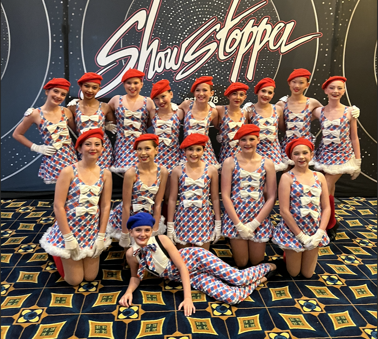 A group of young girl dancers dressed in red, white, and blue checkered costumes with bows and berets, posing for a photo backstage at a show.