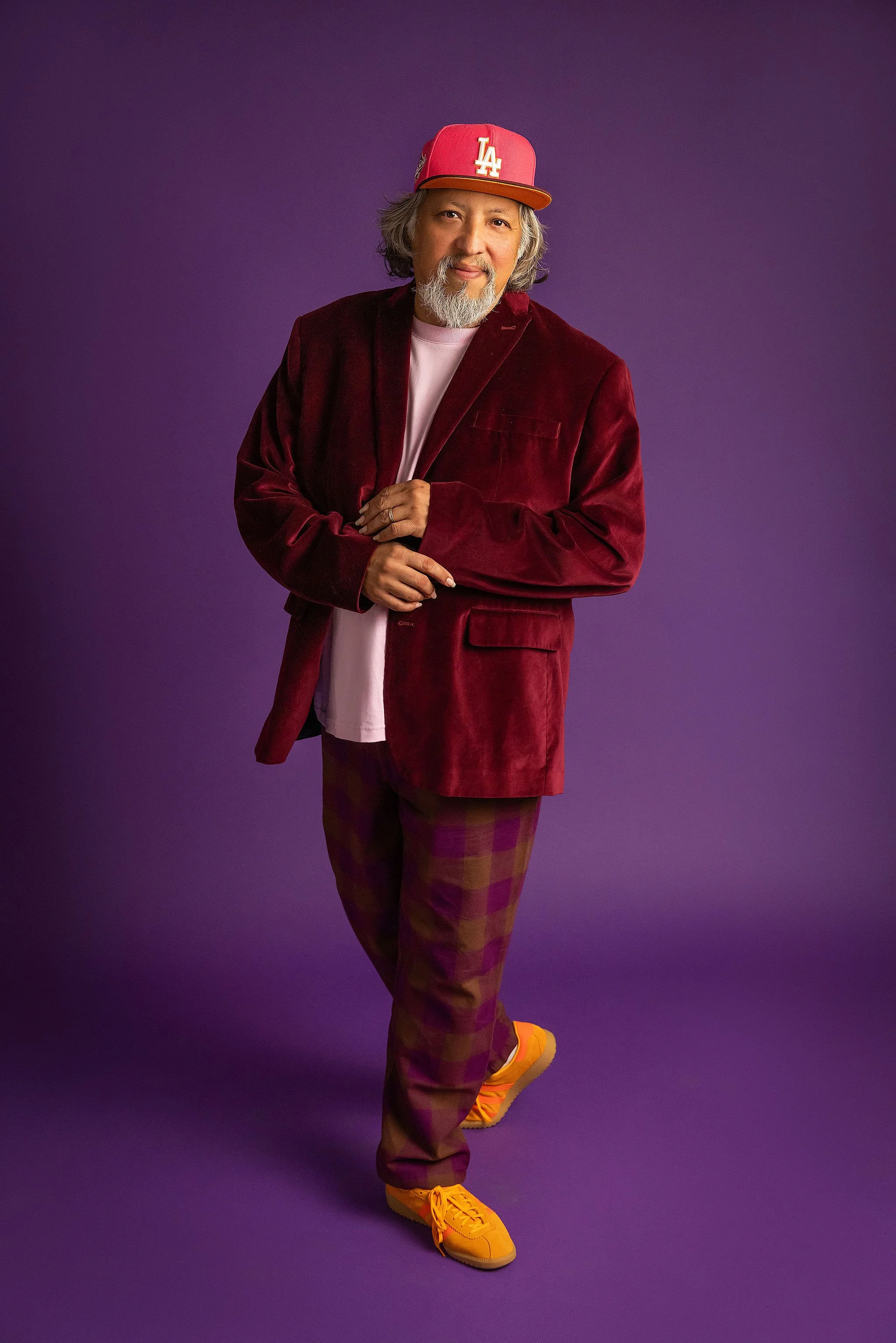 A man with gray hair and a beard smiling, wearing a red LA baseball cap, a dark red velvet blazer, a pink t-shirt, checkered brown and purple pants, and bright yellow sneakers, standing against a purple background.