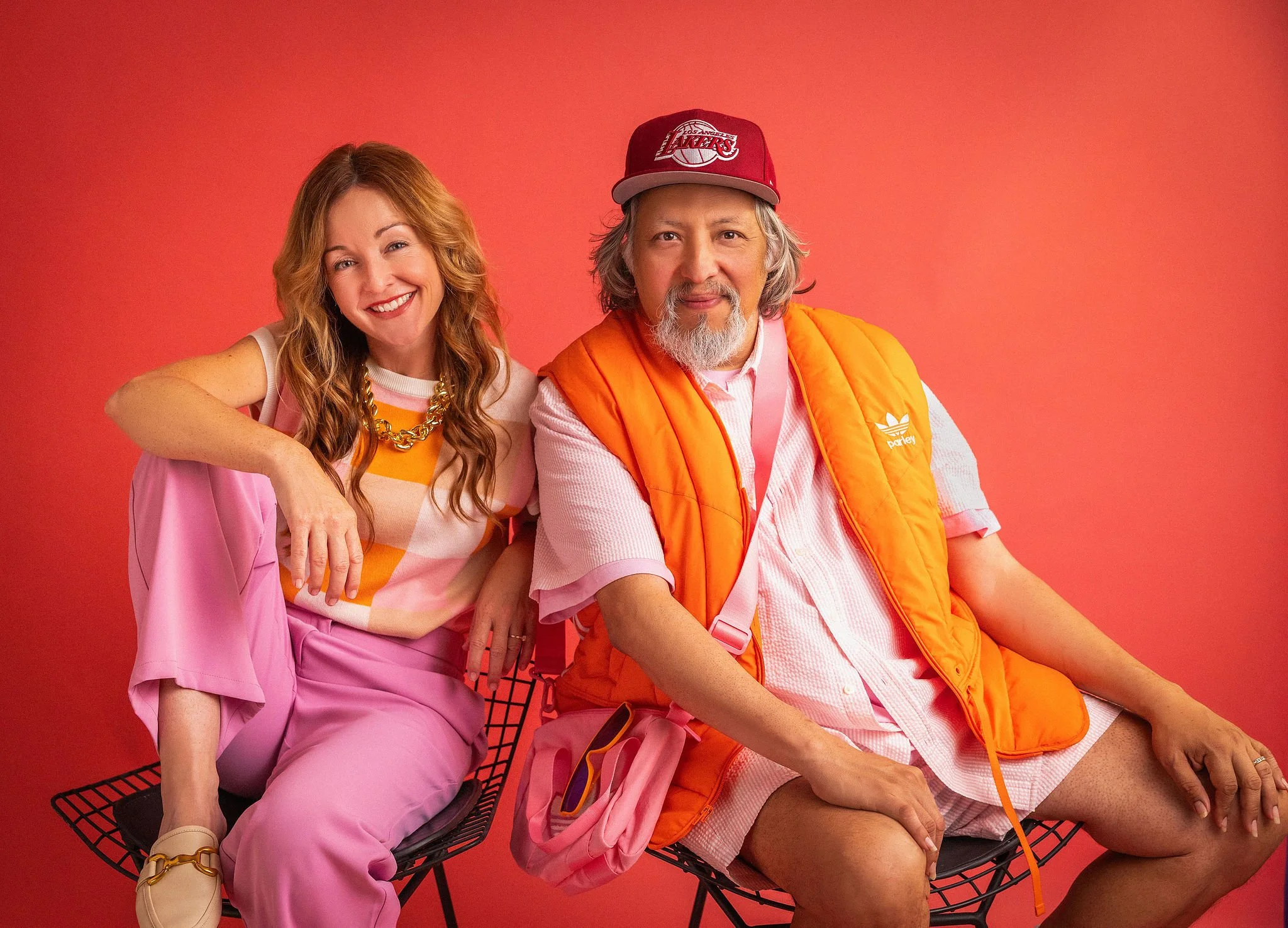 Two people, a woman with wavy red hair and a man with gray hair and beard, sitting on a black wireframe bench against a red background. The woman is smiling, wearing a pink outfit with a white and orange striped shirt and gold jewelry. The man is wearing a red cap, pink shirt, and orange vest, looking directly at the camera.