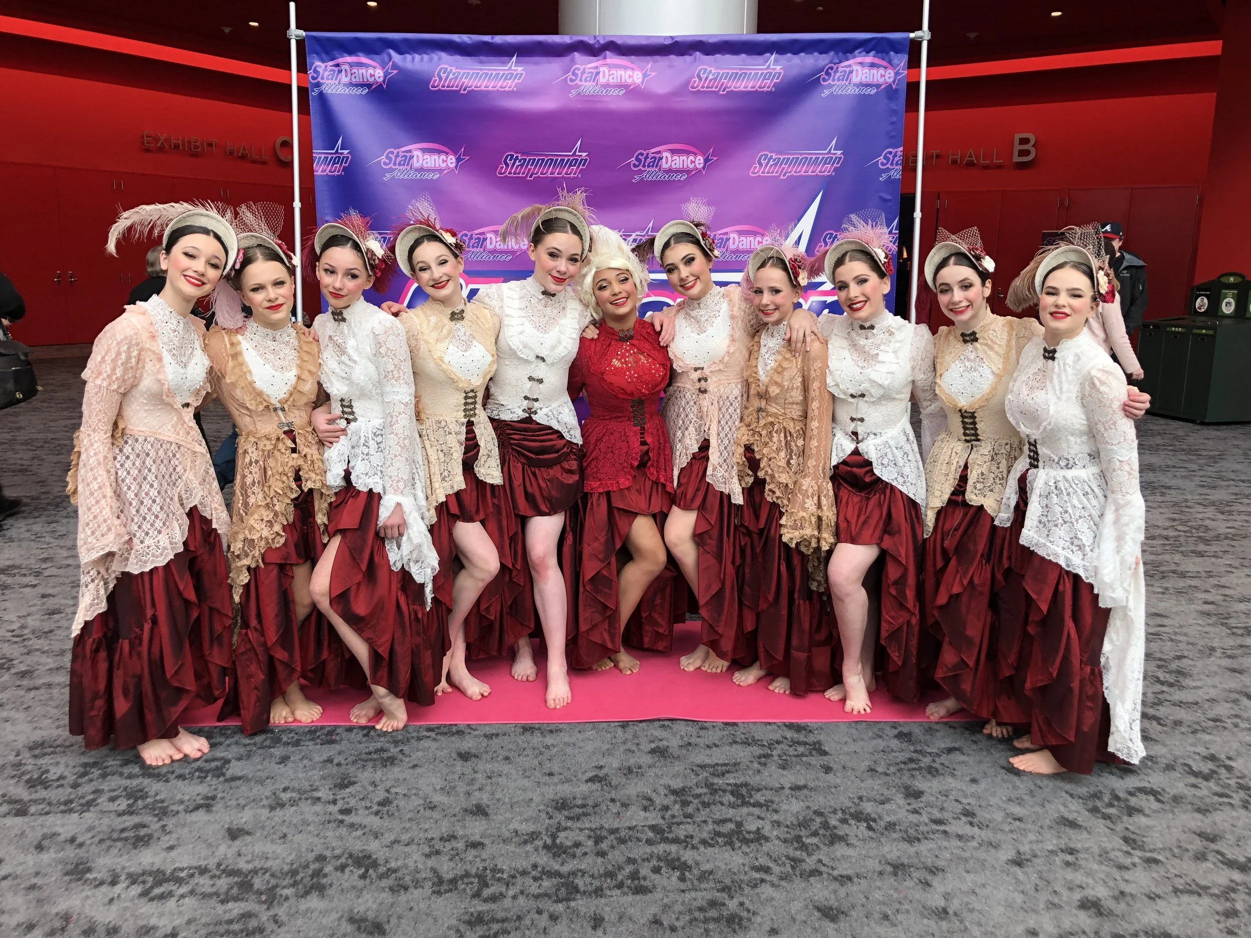 A group of ten young female dancers in vintage-style costumes standing barefoot on a pink carpet in front of a Starpower and Star Dance Alliance backdrop at a dance competition.