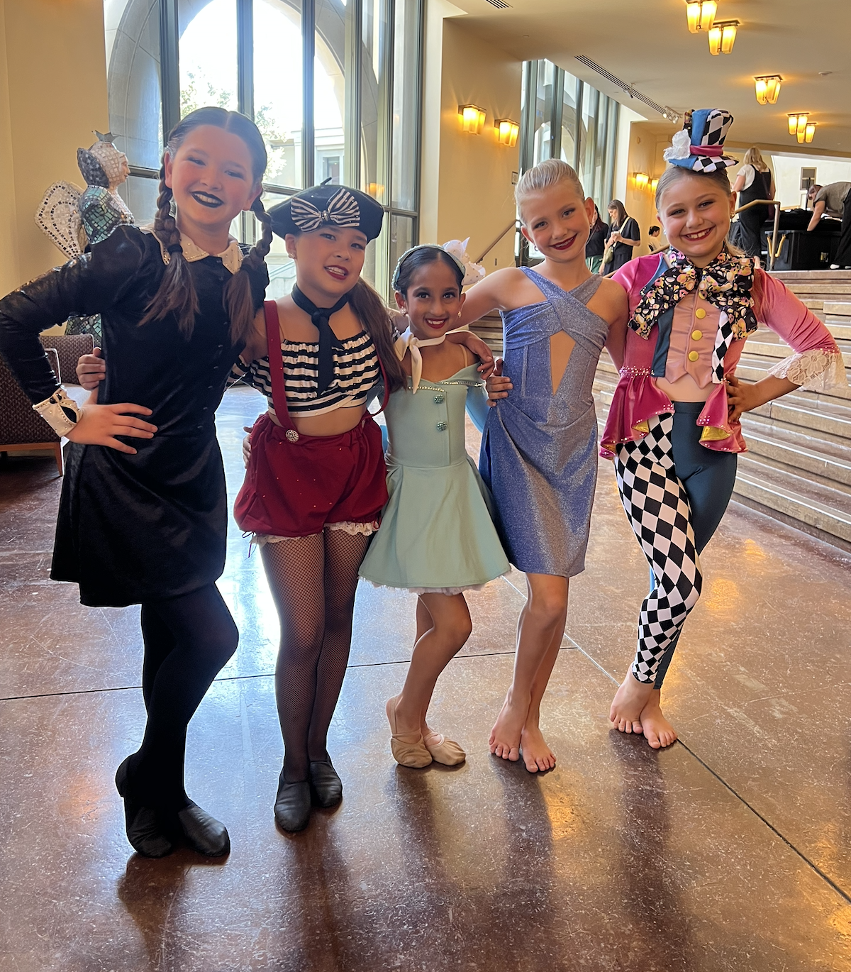 Five young girls dressed in colorful costumes, standing together in a lobby with large windows and staircase in the background.