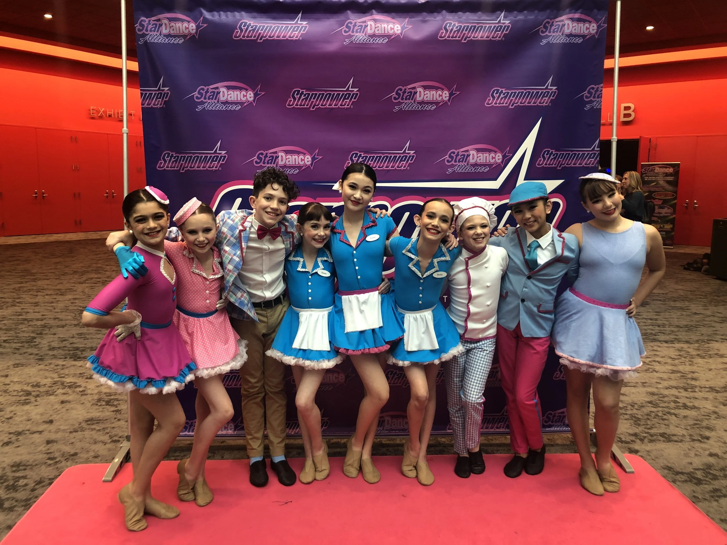 Group of young children wearing colorful costumes, standing on a red carpet in front of a purple backdrop with the 'StarDance Alliance' logo, poses for a group photo.