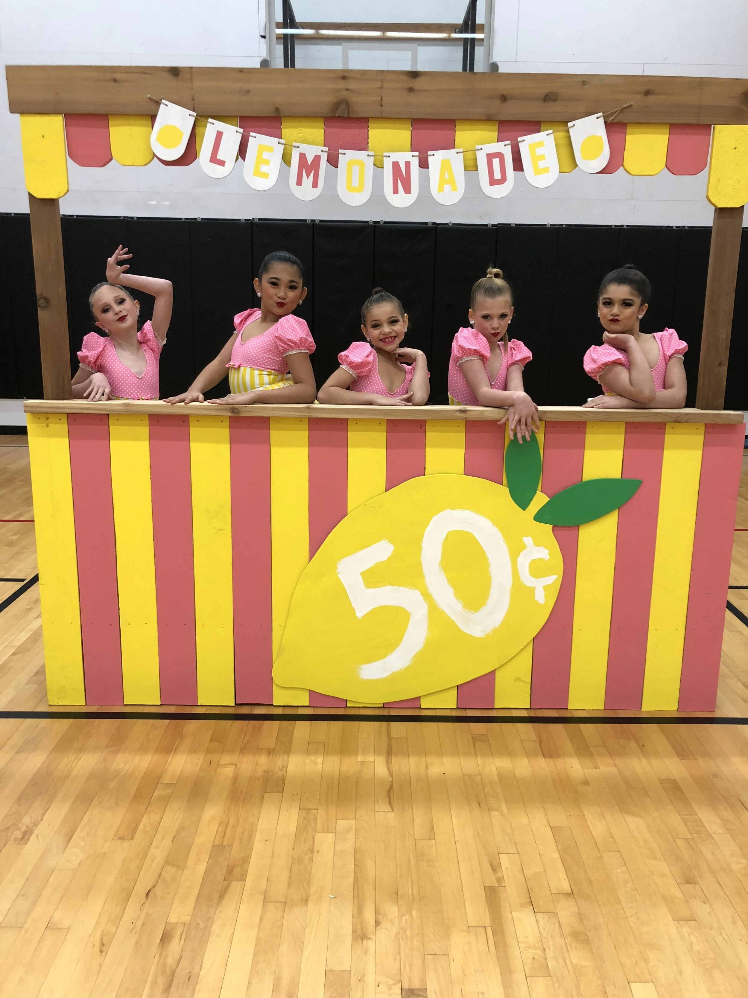 Young girls in pink costumes standing behind a colorful lemonade stand with a yellow lemon graphic and a large sign displaying 50¢.