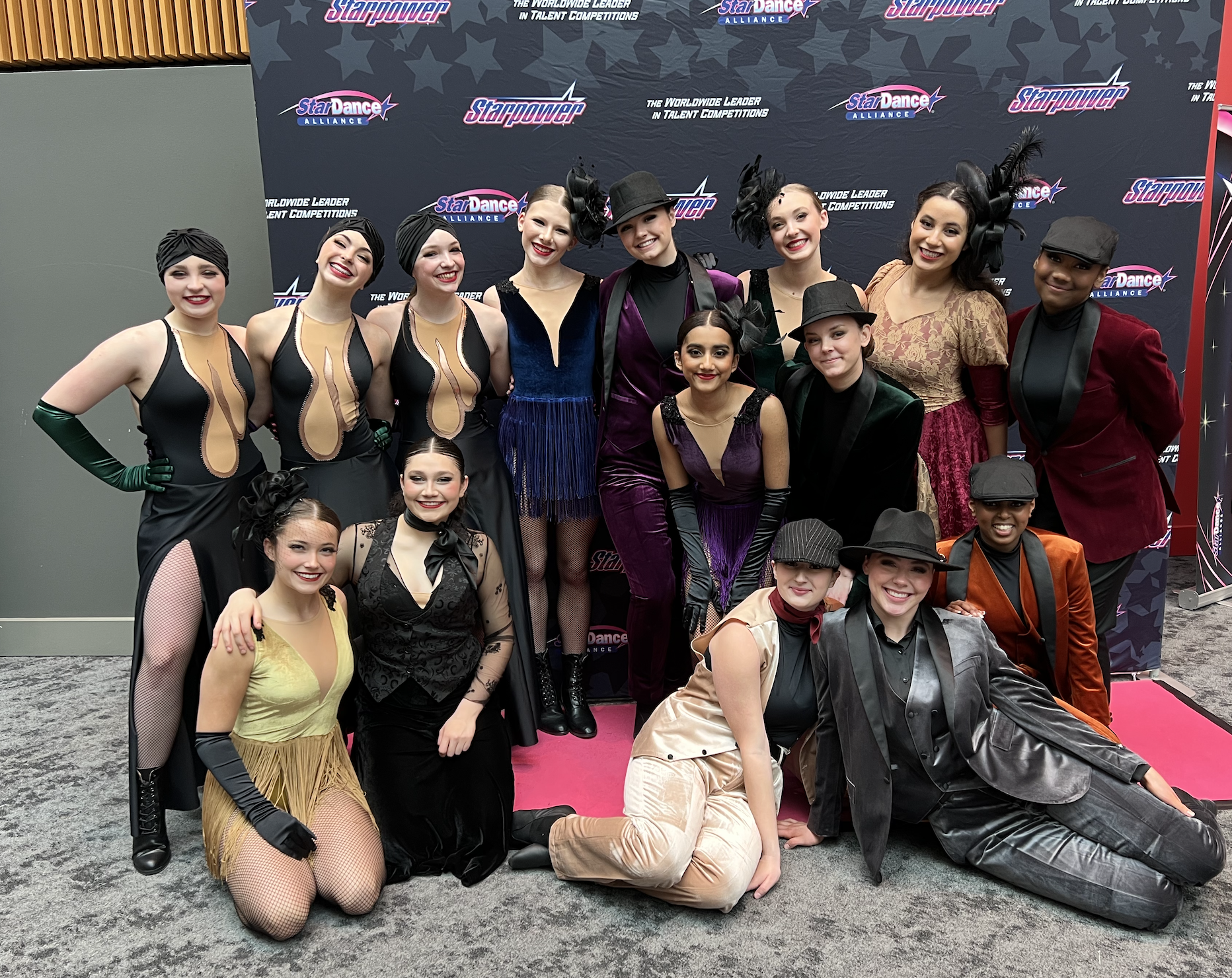 A group of dancers dressed in vintage and theatrical costumes posing at a dance competition, with a backdrop featuring logos of Star Dance Alliance and Starpower.