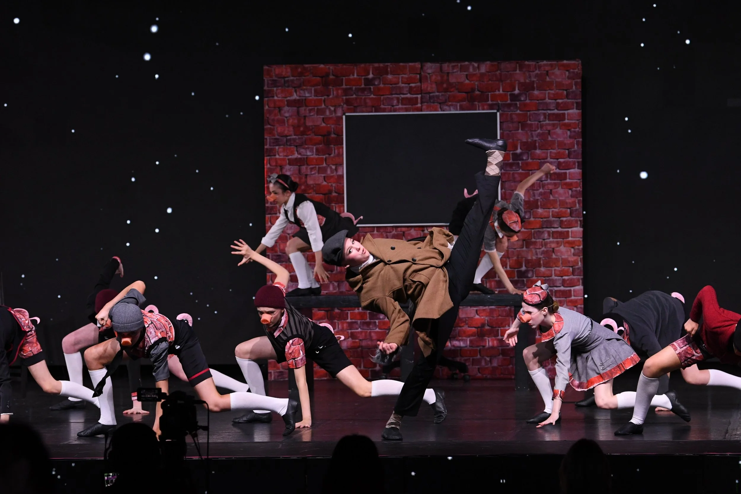 A theater stage with actors in costumes performing a scene. The set includes a red brick wall and a black chalkboard. Actors are dressed as mice with mouse costumes, including ears, noses, and tails, and are in various poses. One actor, dressed as a 
