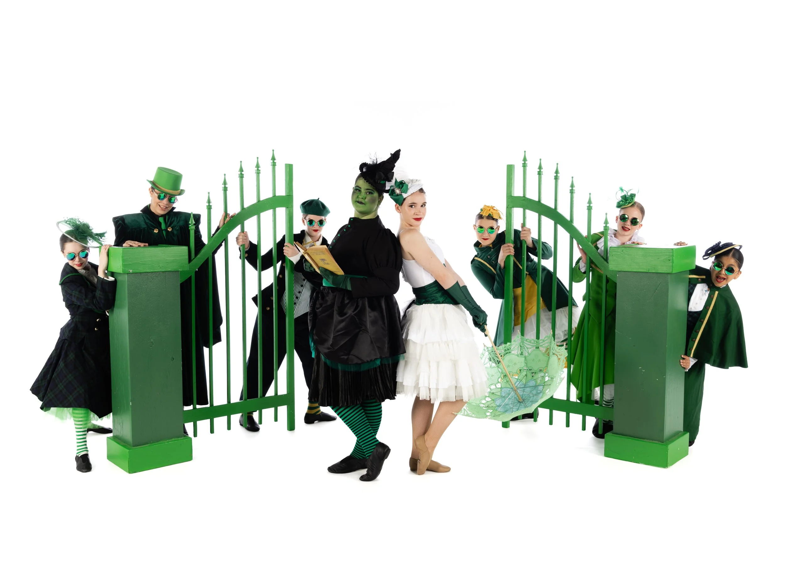 Group of people dressed in costumes with green accents standing around a green gate, some holding umbrellas and wearing sunglasses, on a white background.
