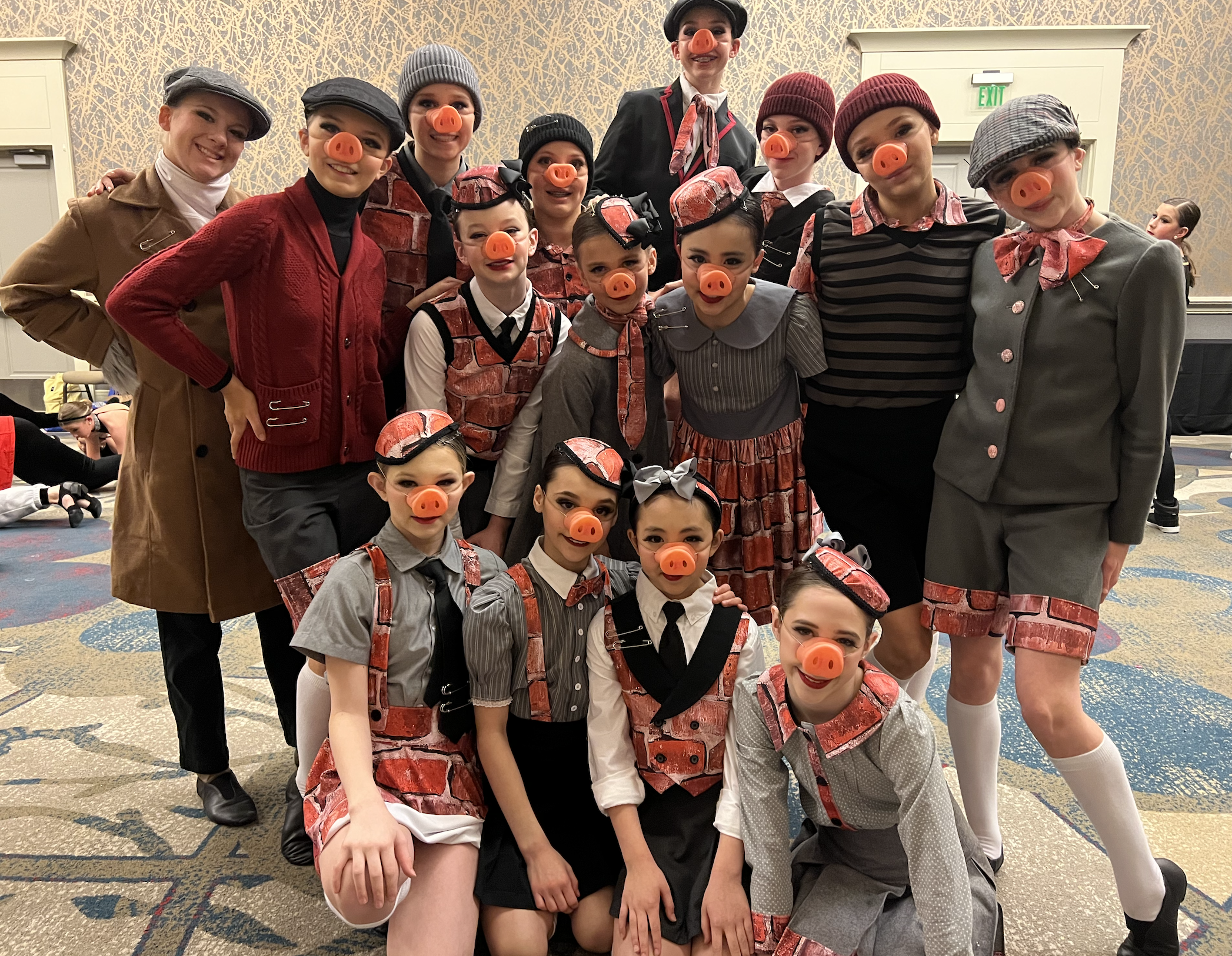 A group of children and two adults dressed in costumes for a pig theme, with pig snout masks and outfits resembling bricks or buildings, posing together indoors.