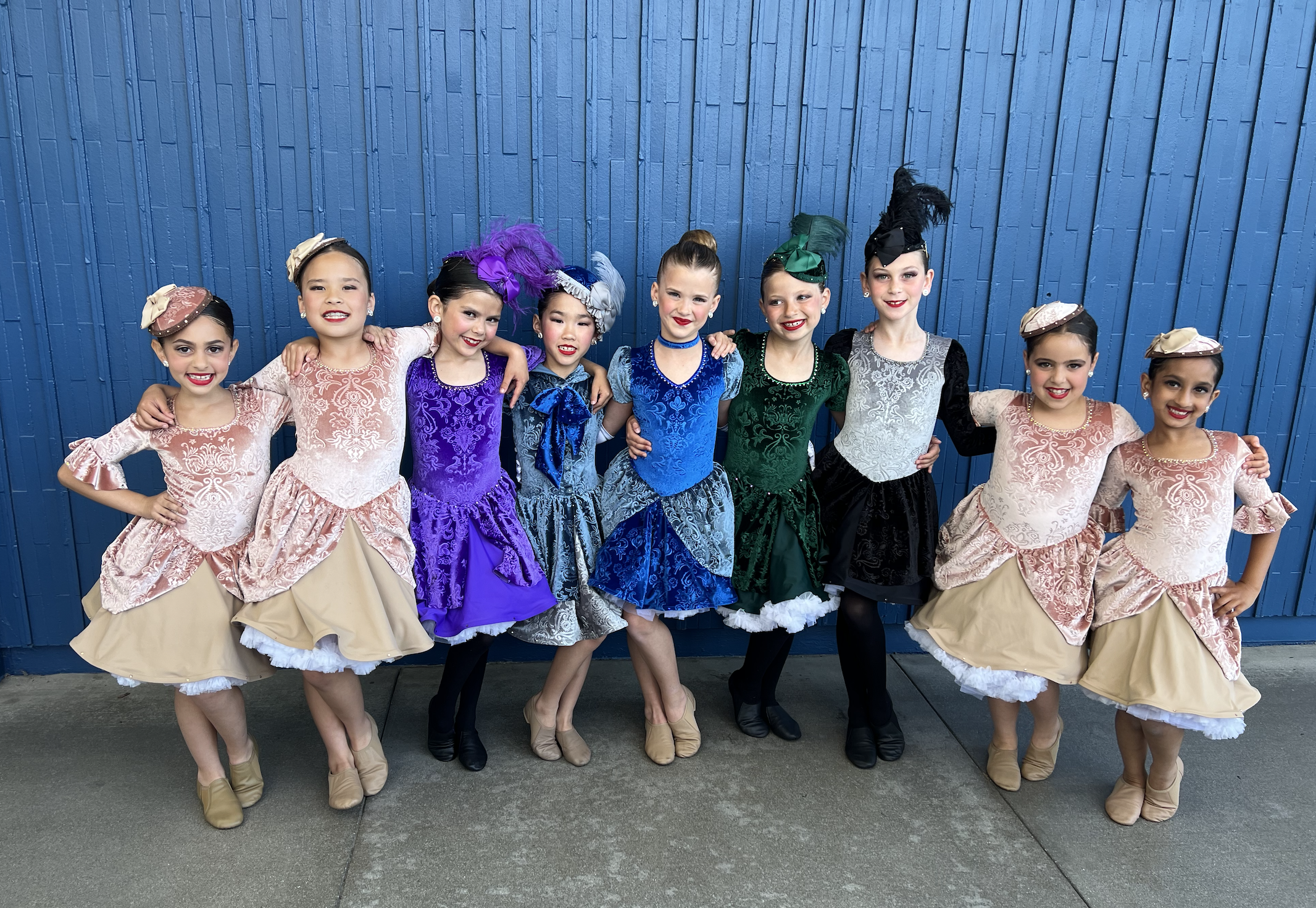 A group of ten girls dressed in colorful, ornate costumes standing together in front of a blue wooden wall. They are smiling and posing with their arms around each other.