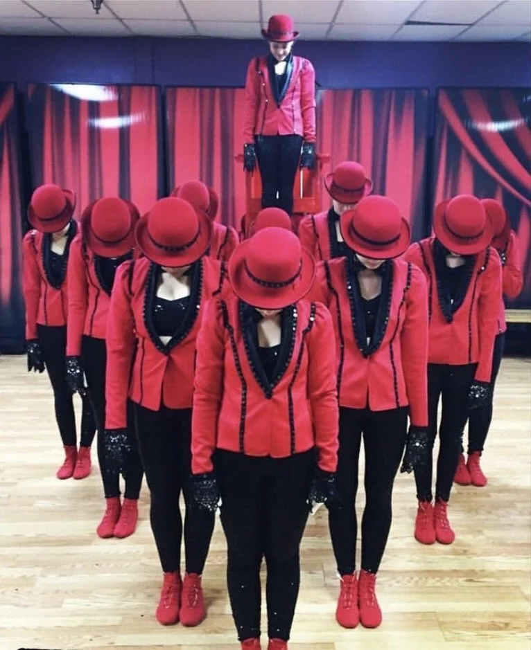 A group of dancers dressed in red jackets, red hats, black pants, and red shoes, performing a synchronized pose on a wooden floor with a red curtain backdrop. One dancer is standing on a platform behind the group.