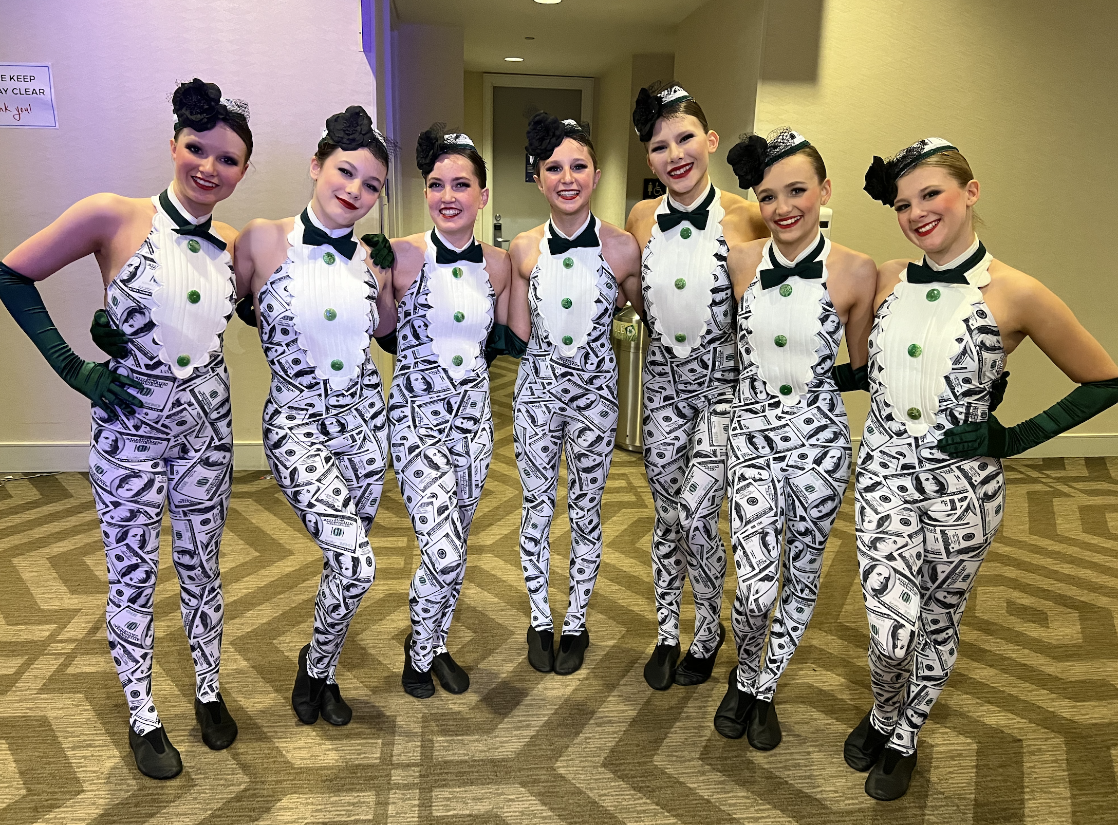 Group of eight women dressed in matching outfits with dollar bill print, black gloves, black shoes, and black headpieces, standing together in an indoor setting.