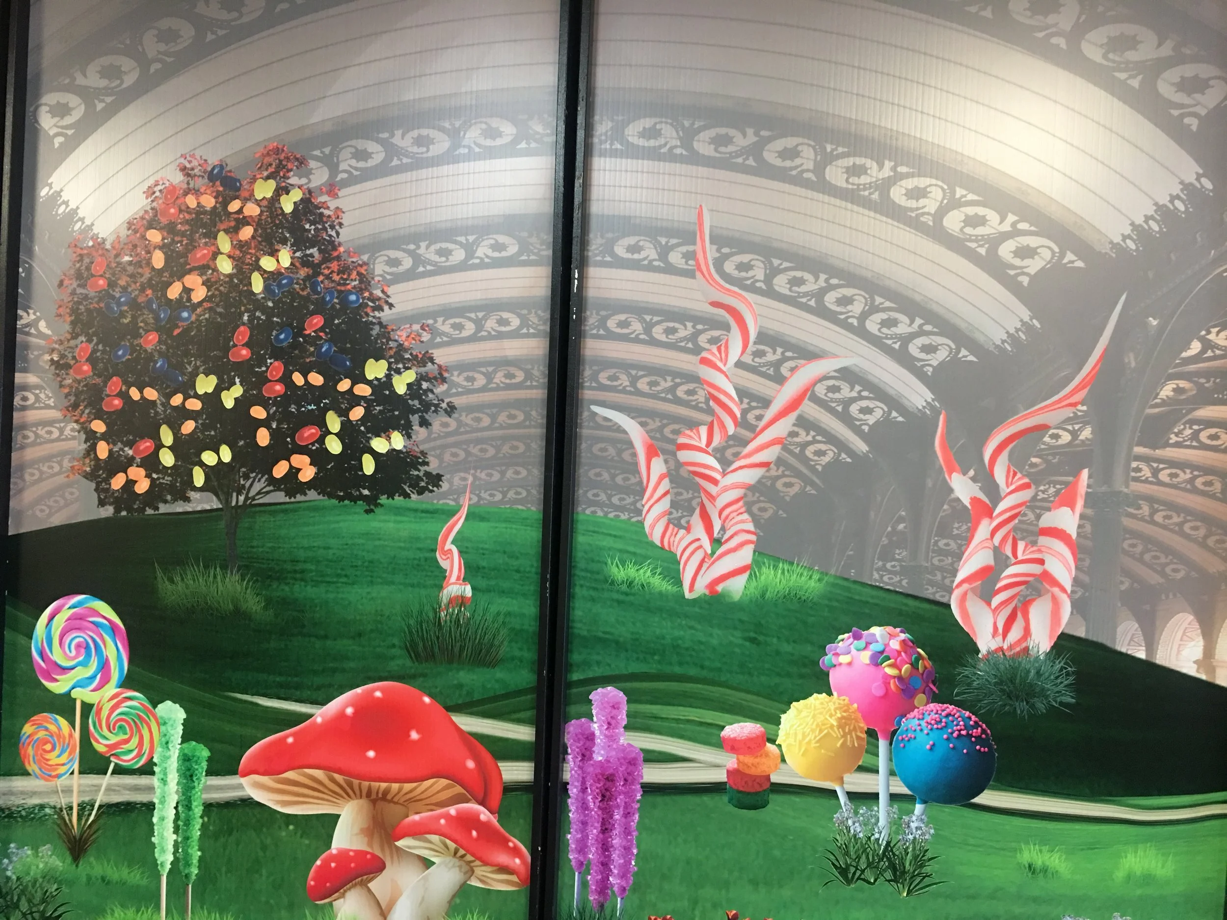 Colorful, whimsical candy-themed scene with a decorated Christmas tree, striped candy canes, large lollipops, and colorful sweets on artificial grass, set against a backdrop of ornate architectural design.