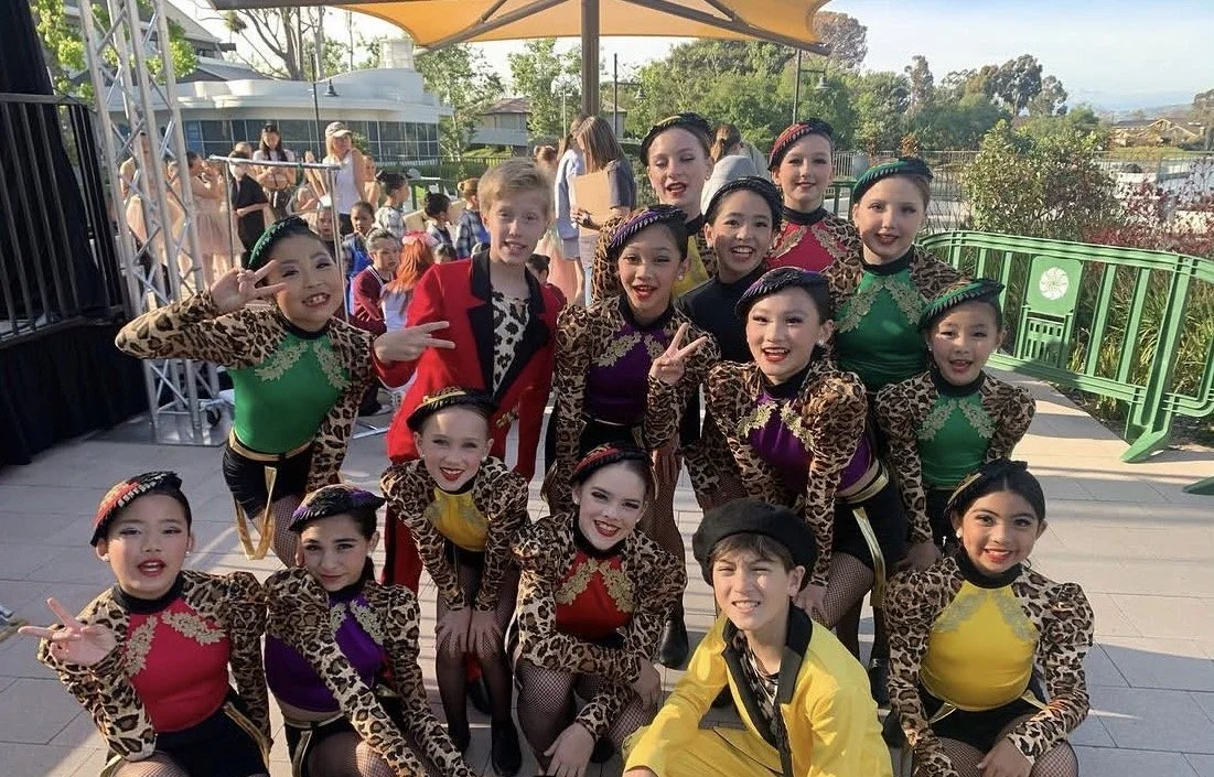 A group of young dancers in leopard print costumes, posing and smiling outdoors at a dance event, with some making peace signs.