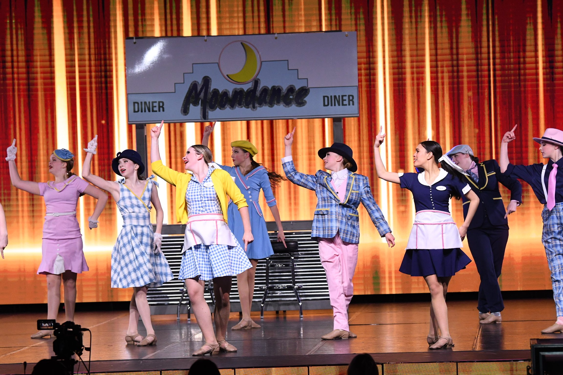 A group of women dressed in vintage 1950s-style clothing performing a musical on stage, with a large sign that reads 'Moondance Diner' and a moon logo in the background.