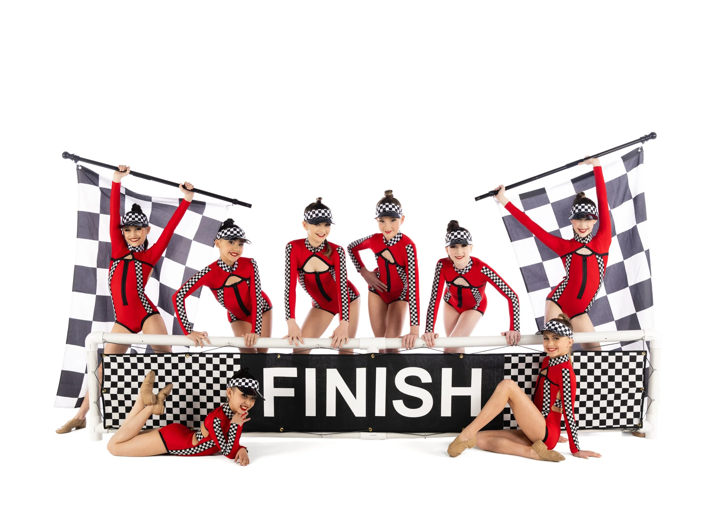 Group of female performers in red and black checkered racing costumes celebrating at a finish line, holding checkered flags, with some lying or sitting and cheering in front.