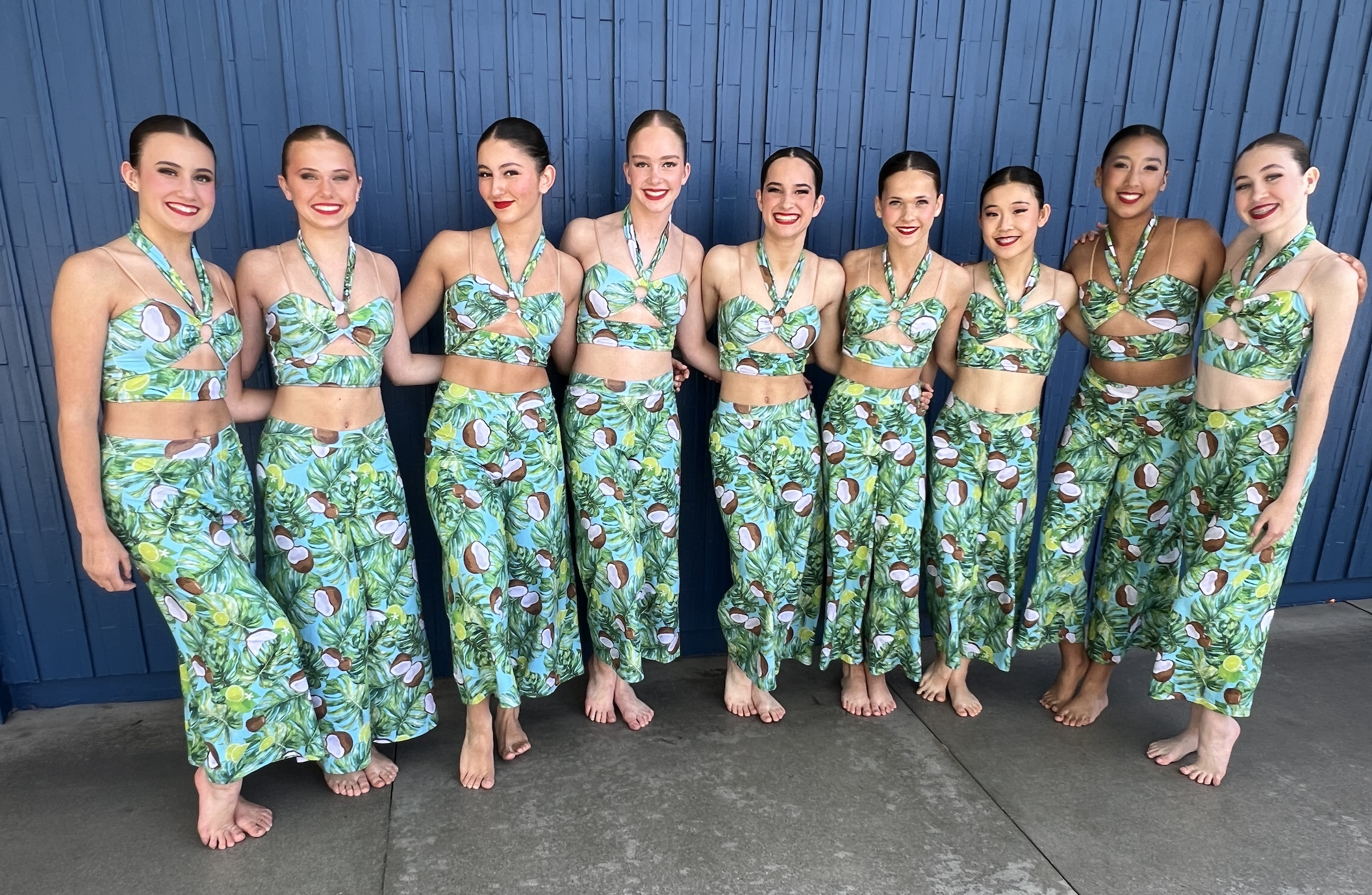 Eight women in matching tropical-themed dance costumes standing barefoot against a blue wooden wall, smiling at the camera.