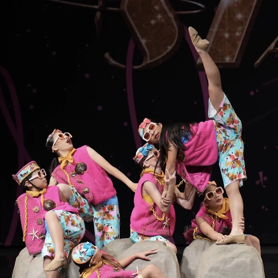 Children dressed in colorful, playful costumes performing on stage. They are wearing bright pink vests, patterned pants, sunglasses, and accessories like chains and hats. The scene appears lively and whimsical, likely part of a theatrical or dance pe
