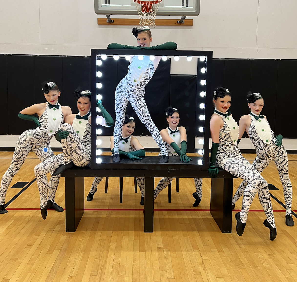 Group of six women dressed in cash-themed costumes posing around a mirror with lights, in a gymnasium with a basketball hoop in the background. One woman stands inside the mirror frame, while the others pose around it.