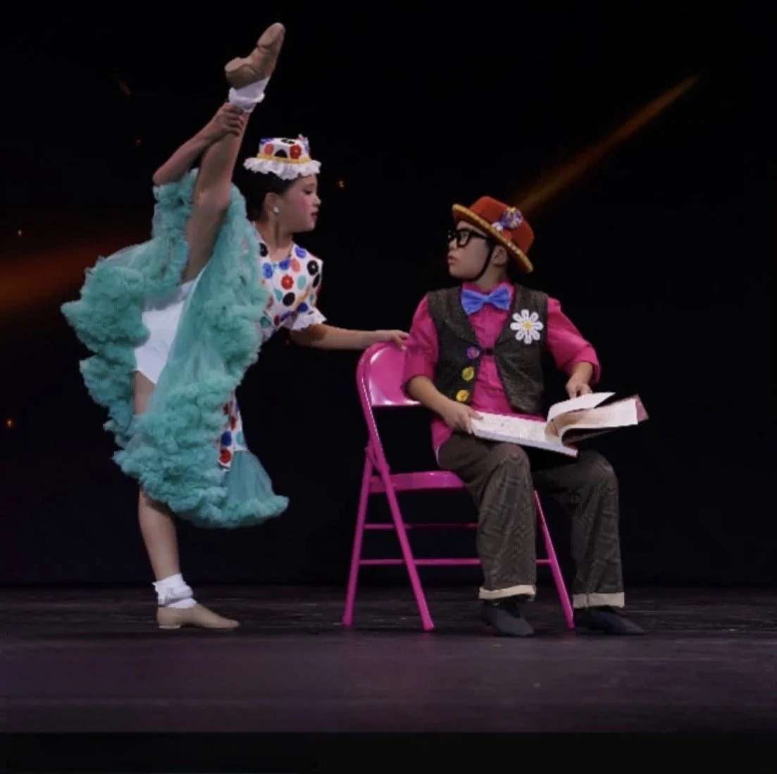 Two children performing in a theatrical play. One girl dressed in a colorful polka dot dress and a hat with laces is lifting the other girl, who is sitting on a pink chair. The girl in the chair is wearing glasses, a red hat, a pink shirt, a vest with a flower on it, and is holding a book.