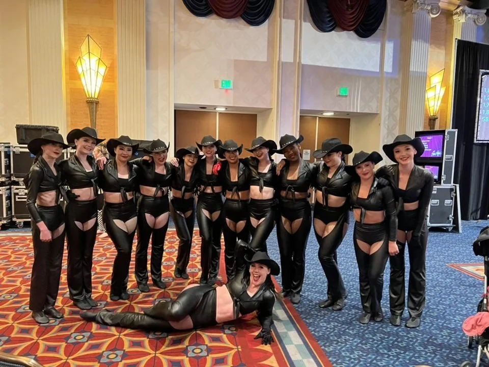 A group of 13 women in black leather cowgirl costumes, with one lying on the floor in front, posing for a photo in a ballroom.