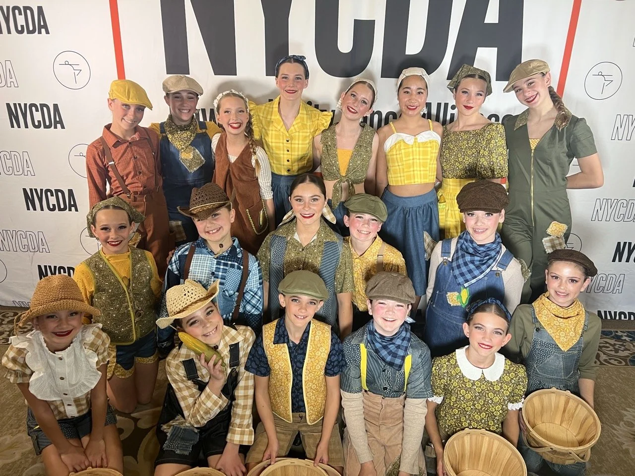 A group of young dancers in vintage-inspired costumes pose in front of a NYCD logo backdrop, with some holding baskets.