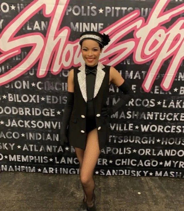 A woman in a black and silver showgirl costume with fishnet stockings standing in front of a backdrop with the words 'Showstopper' and a list of city names.
