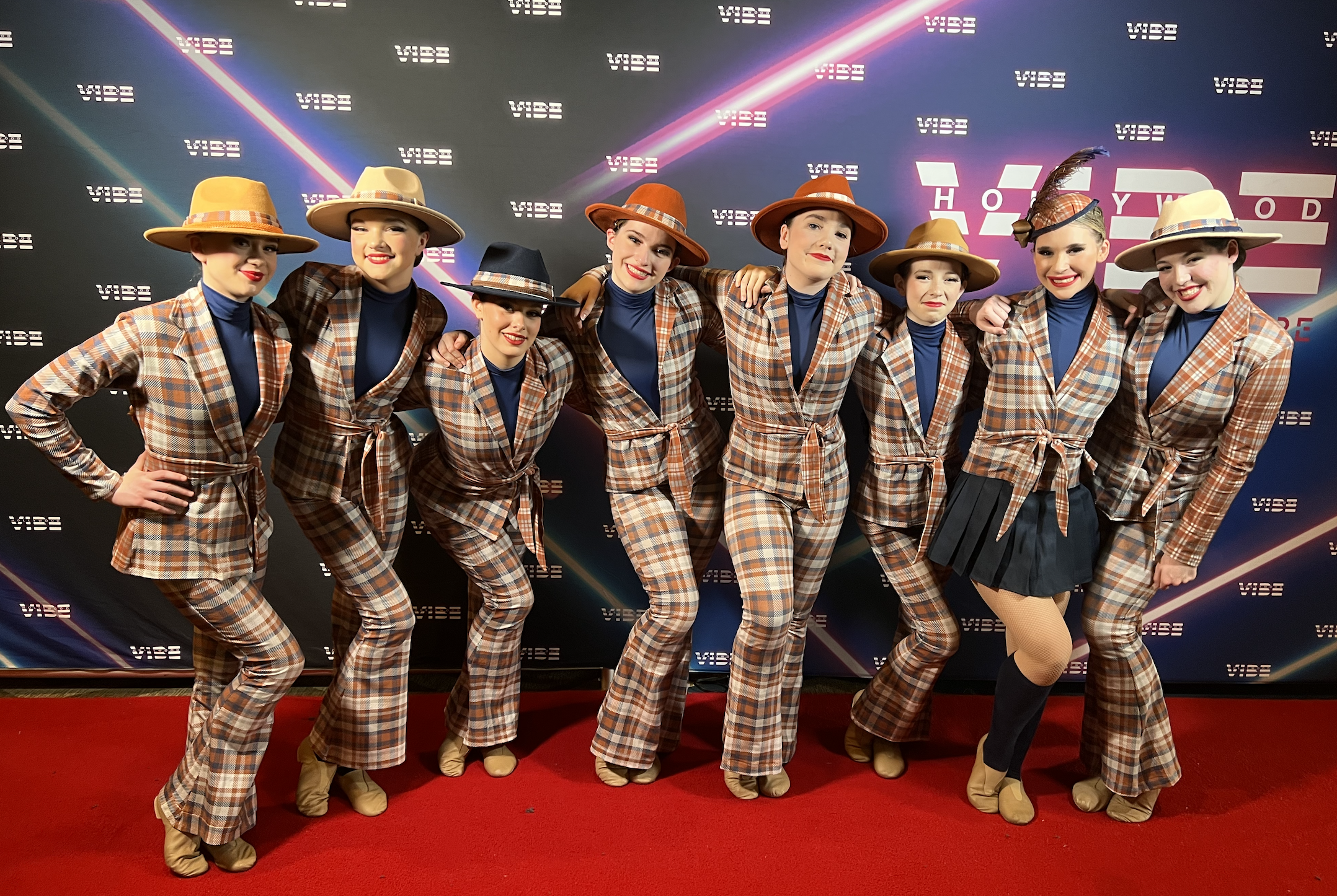 Group of ten women dressed in matching plaid costumes with hats, posing on a red carpet at an event, with a backdrop displaying the WIBE logo and neon lights.