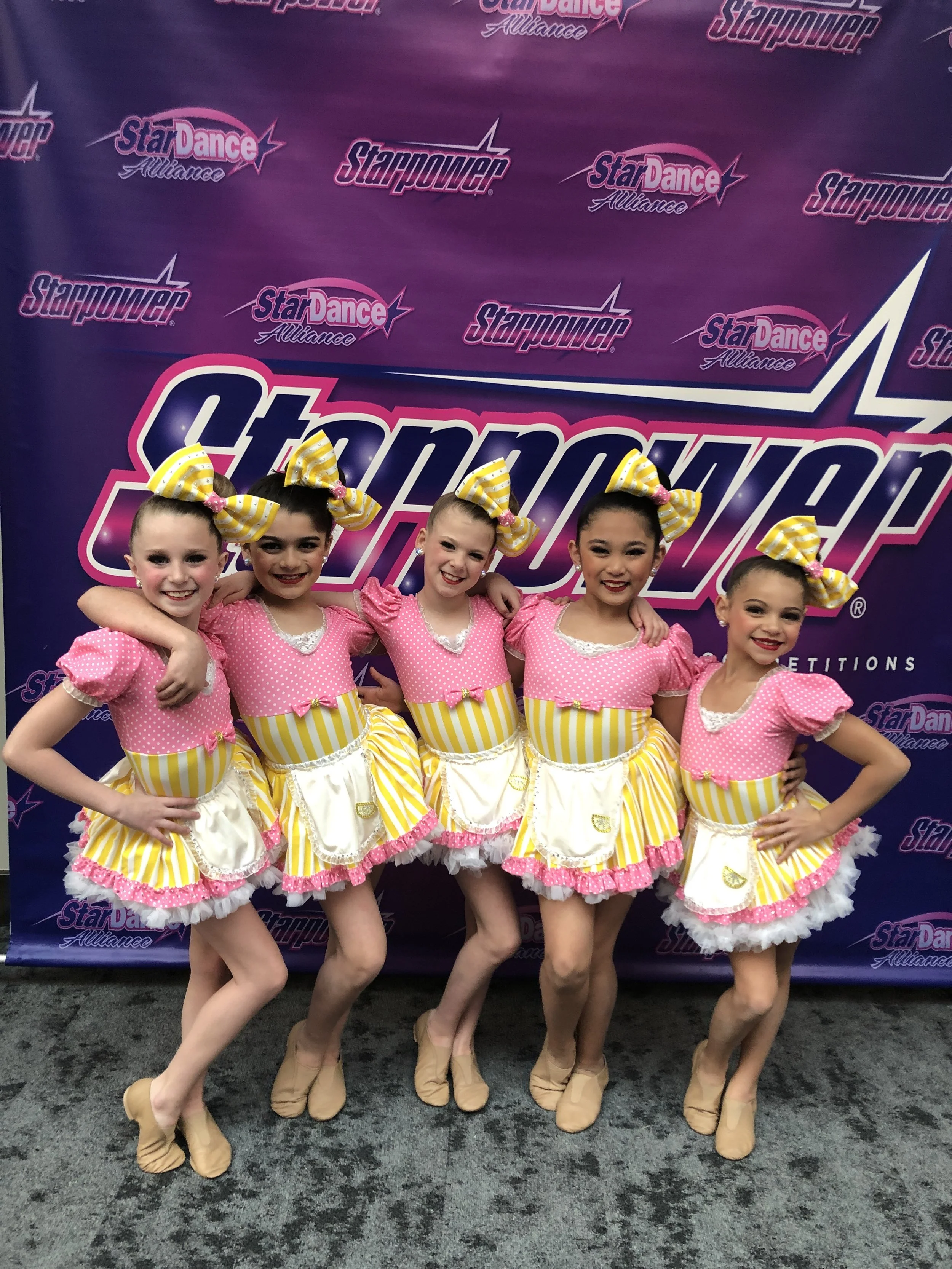 Five young girls dressed in pink polka dot costumes with yellow and white striped bows in their hair, posing together at a dance competition in front of a purple backdrop with the logos 'StarDance Alliance' and 'StarPower.'