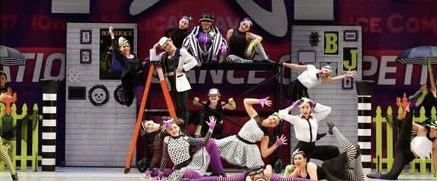 Group of performers dressed in colorful costumes posing on and around a staged set with a ladder, props, and backdrop with signs, in a dance or theatrical performance.