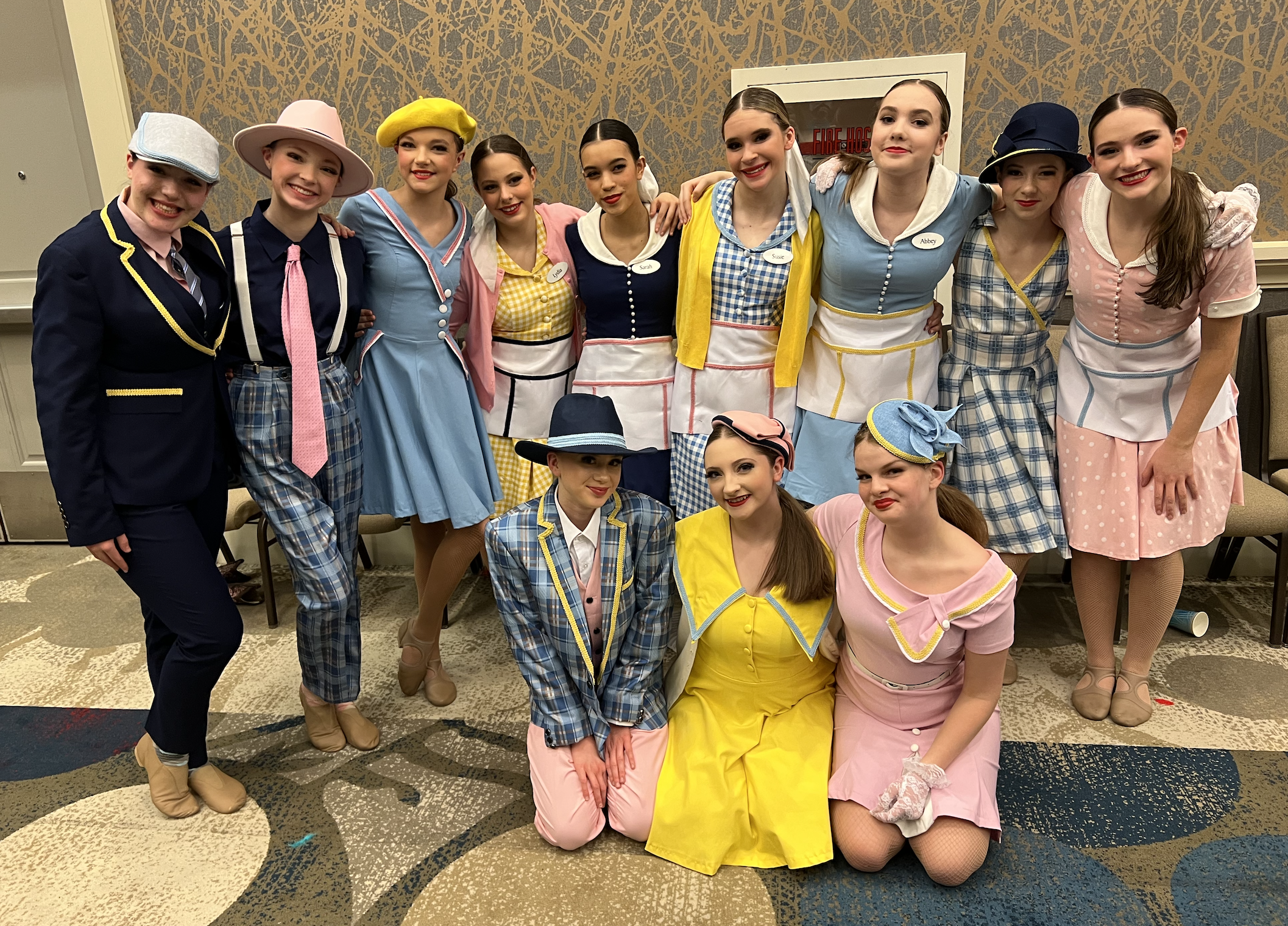A group of young women dressed in vintage-style dresses with aprons, hats, and makeup, posing together in an indoor setting, smiling at the camera.