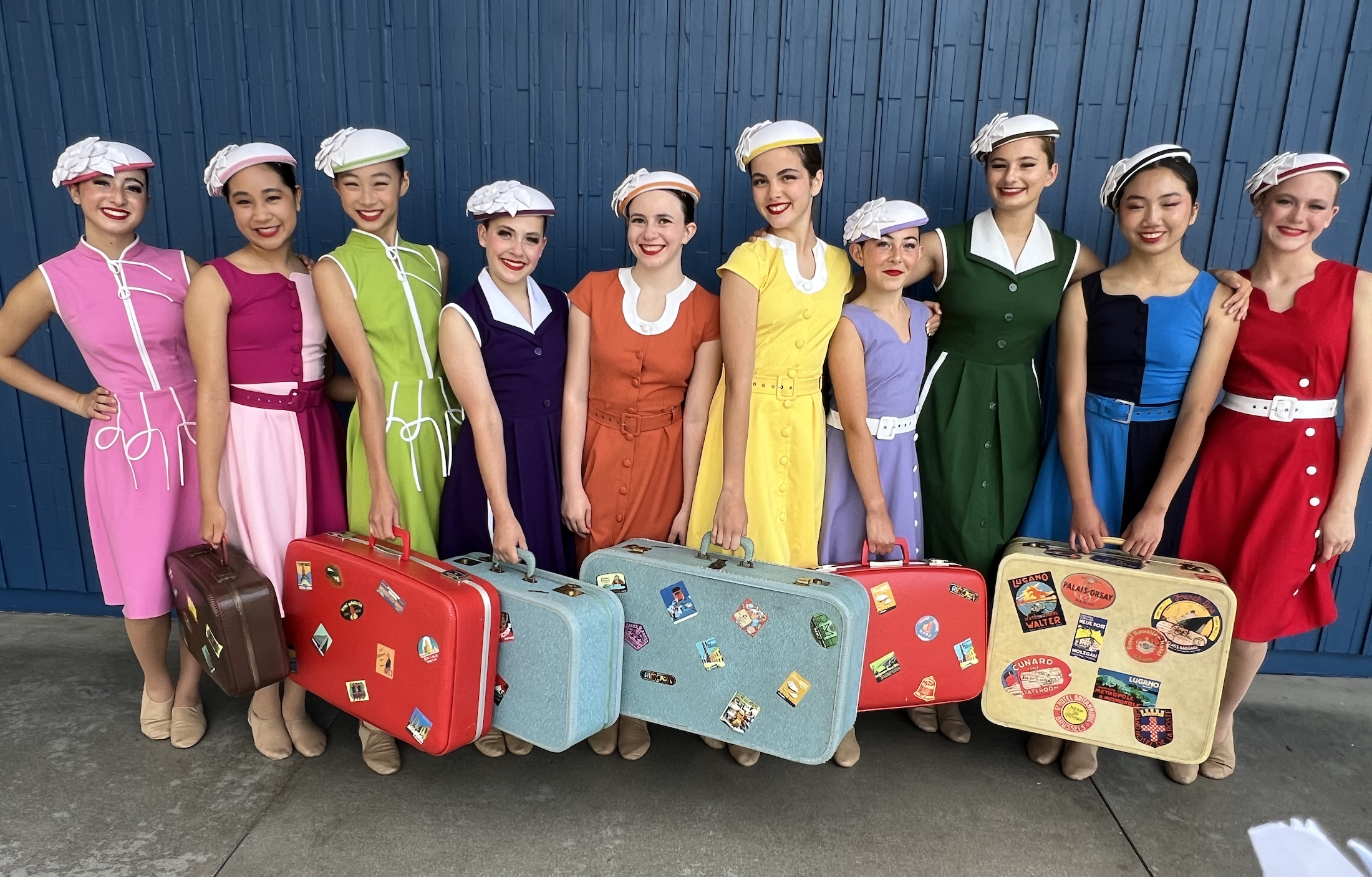 A group of ten women dressed as vintage flight attendants in colorful dresses and matching hats, standing in front of a blue wall, holding vintage suitcases with travel stickers.