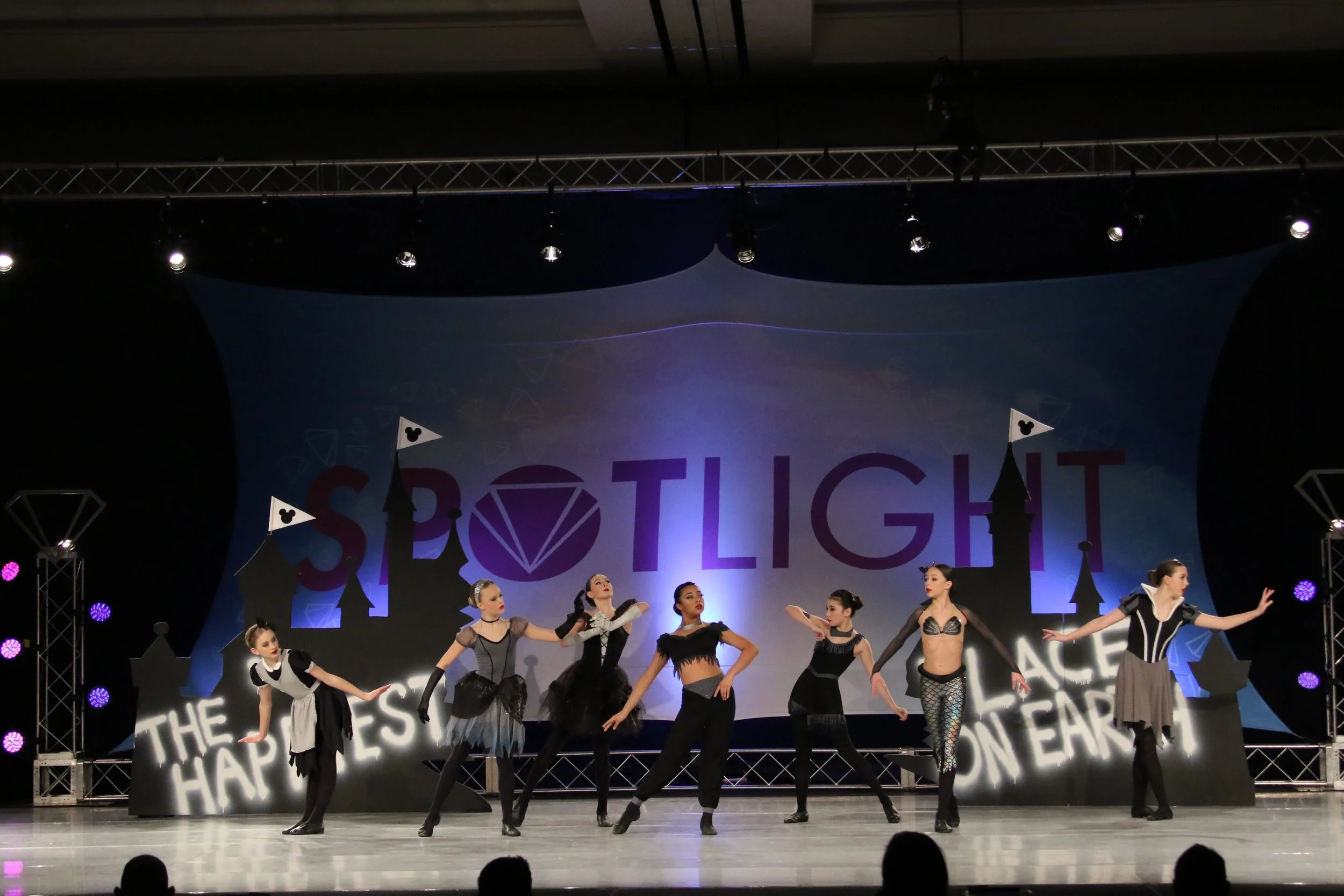 Dance performance on stage with young female dancers in costumes, backdrop displays the word 'SPOTLIGHT' with castle and diamond decorations, stage lights and audience in foreground.
