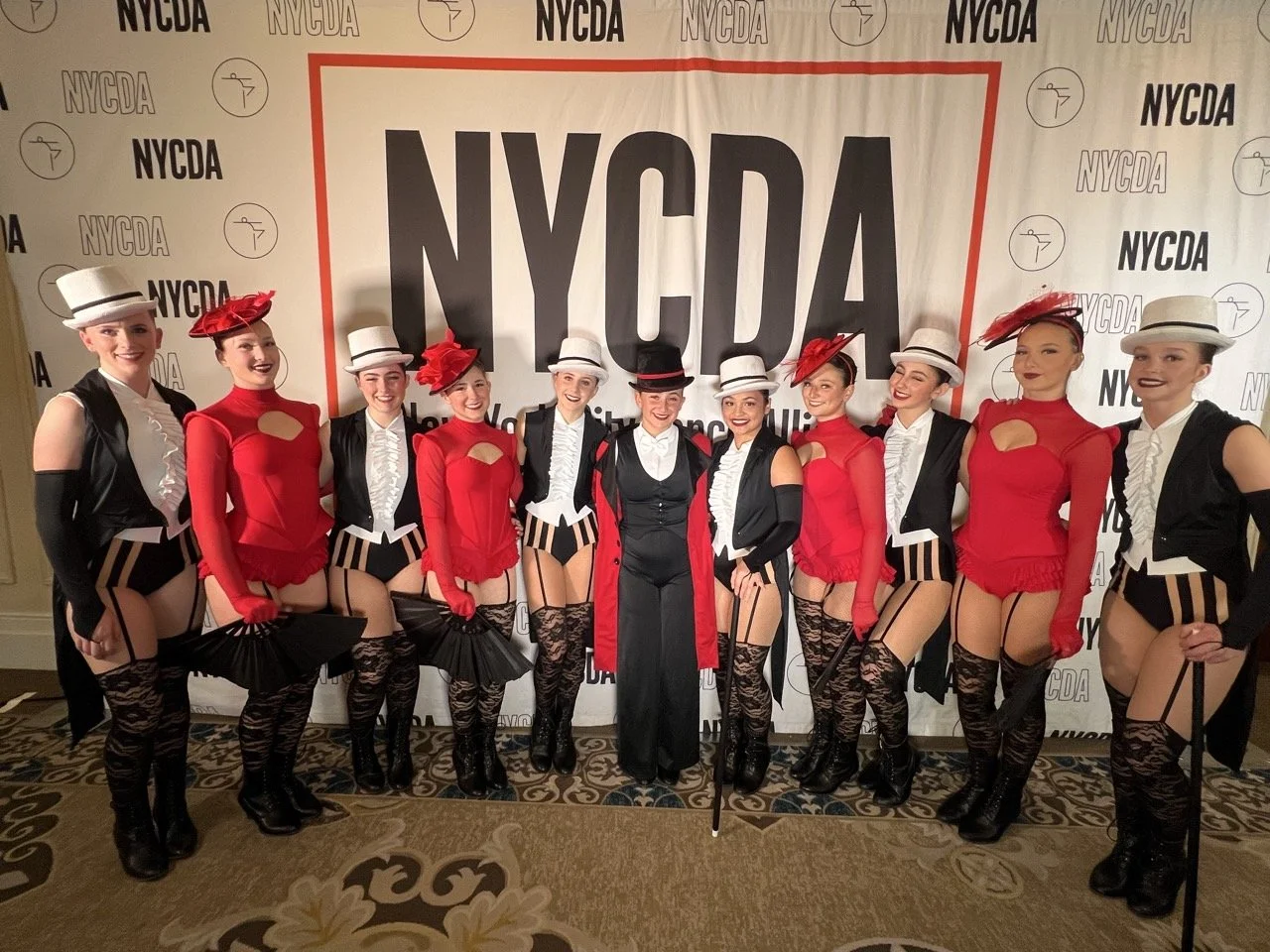 Group of performers in vintage cabaret costumes at NYCDA event, wearing top hats, corsets, striped tights, and holding fans, posing in front of a NYCDA backdrop.