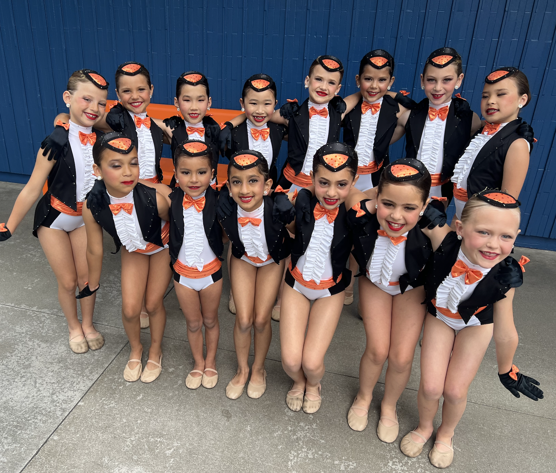 A group of young girls in dance costumes pose together for a photo. They are wearing matching black, orange, and white costumes with orange bow ties and beige ballet shoes. Some girls are smiling, and they are standing in front of a blue wall.