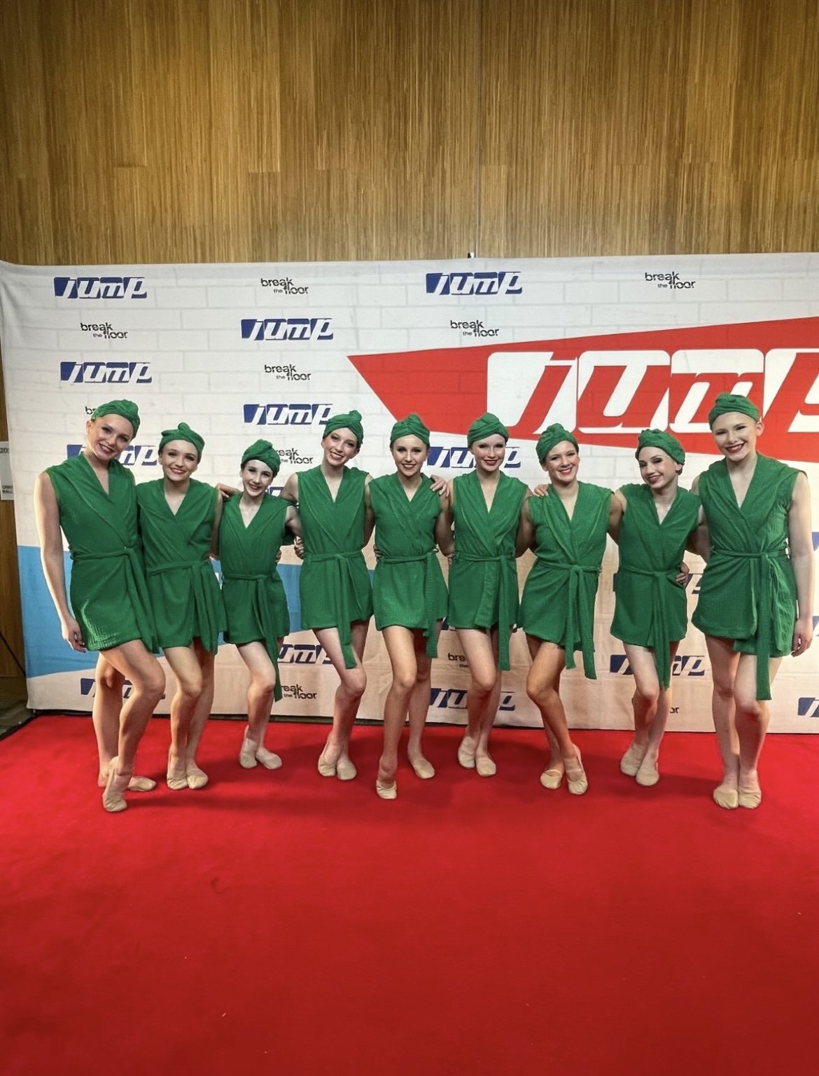 Group of nine female dancers dressed in green costumes and matching headscarves, posing with arms around each other on red carpet at a dance or performance event.